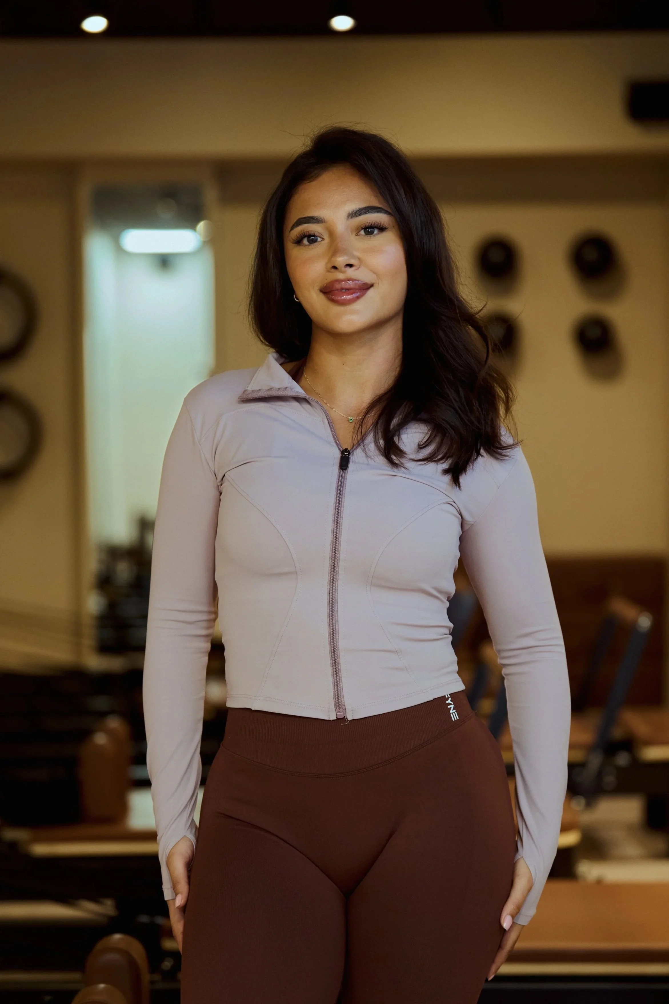 Pilates instructor bij Pilates Avenue in Groningen in een taupe rib sportset, glimlachend in de warme studio setting.