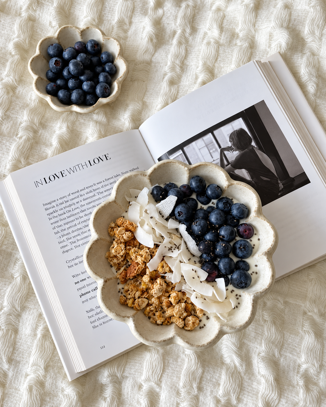 Open book with black and white photo, two ceramic bowls filled with blueberries, granola, coconut flakes, and chia seeds, on a textured cream-colored fabric surface.