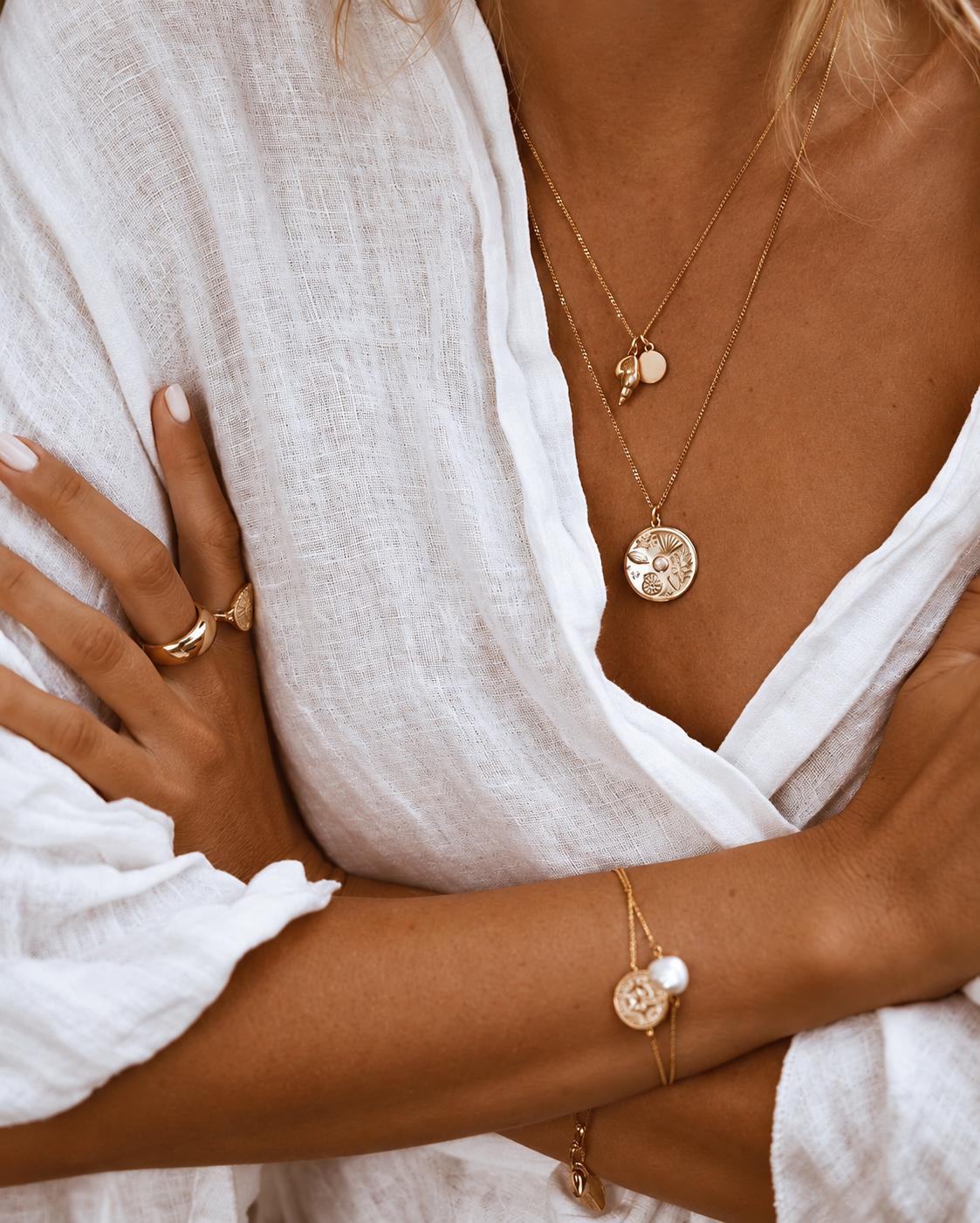 Close-up of a woman wearing layered gold necklaces, including a round pendant with engraved nature images, a smaller pendant, and a trio of small charms, as well as a gold ring and bracelet, all paired with a white linen outfit.
