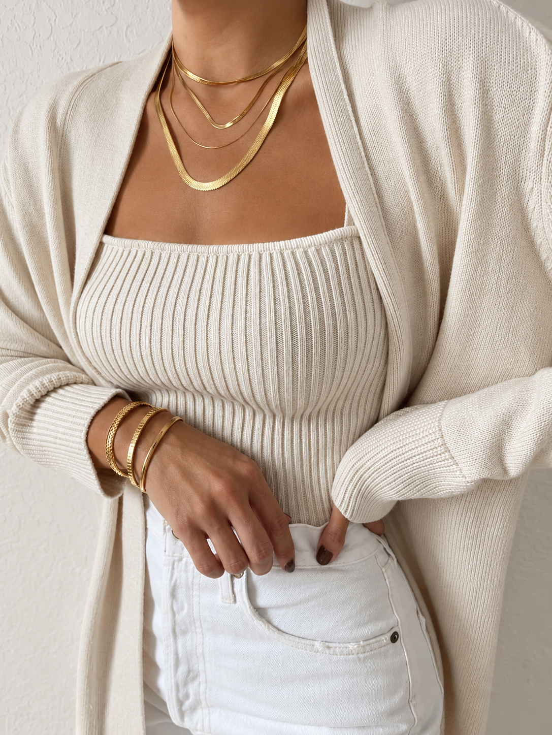 Close-up of a woman wearing layered gold necklaces, multiple gold bracelets, and a cream-colored, ribbed tank top with a matching cardigan, paired with white jeans.