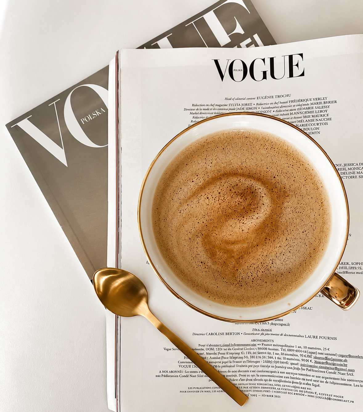 A cup of frothy coffee with a gold rim and handle, resting on a newspaper with Vogue magazine, gold spoon, and another magazine in the background.