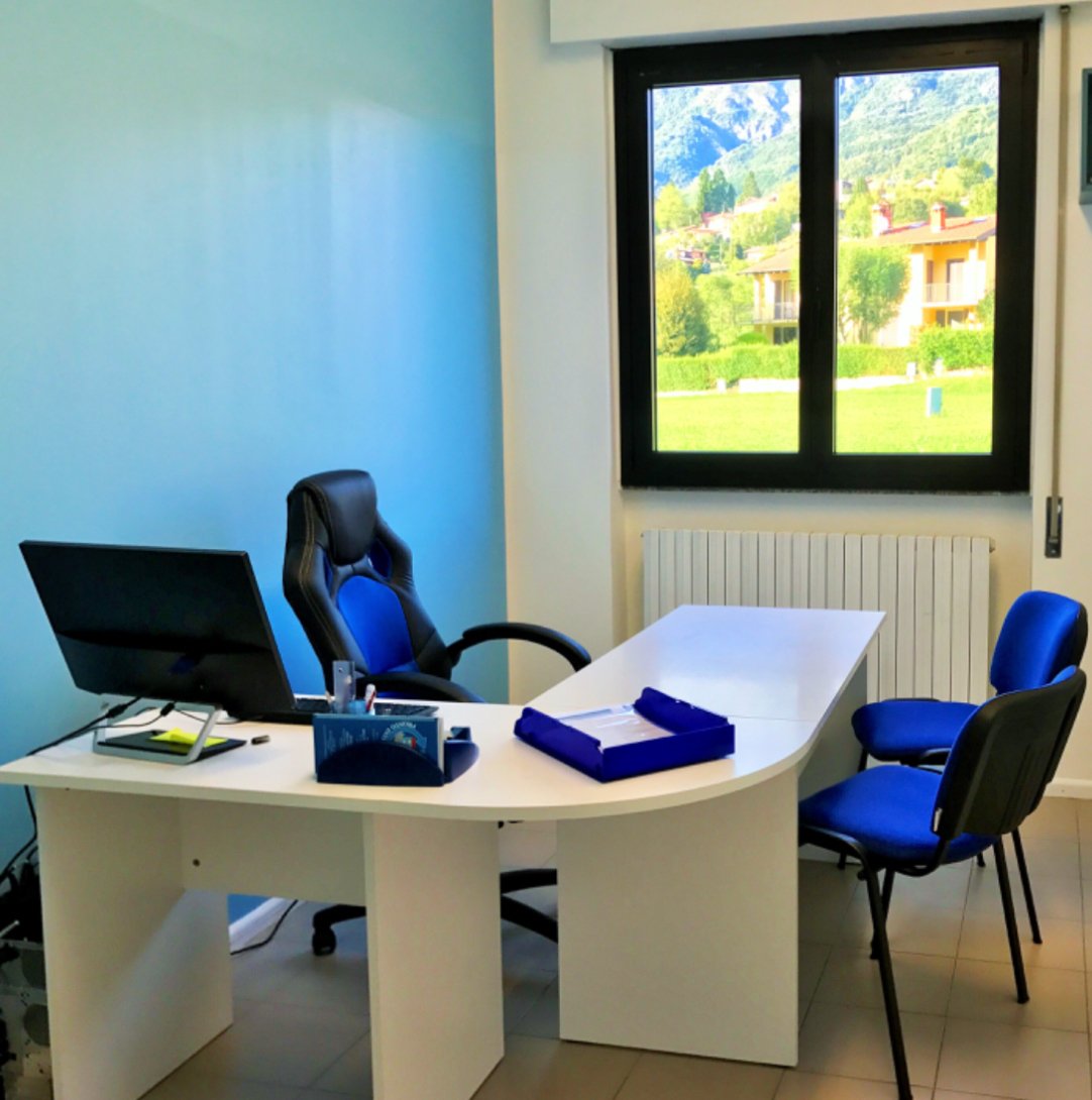 Ufficio moderno con scrivania bianca, sedia da ufficio nera e blu, computer, finestra con vista su paesaggio verde.