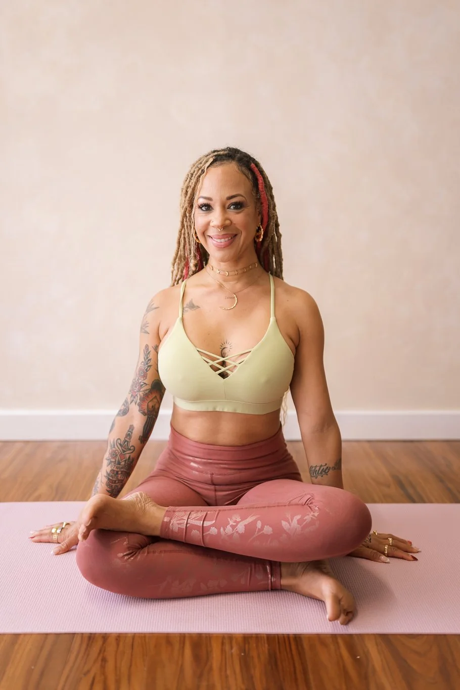 Woman with tattoos in yoga attire sitting on a mat in a yoga pose, smiling at the camera.