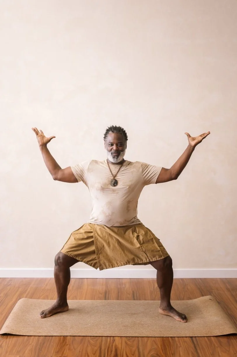 Man doing a yoga pose on a mat, wearing a beige shirt and brown skirt, wooden floor background.