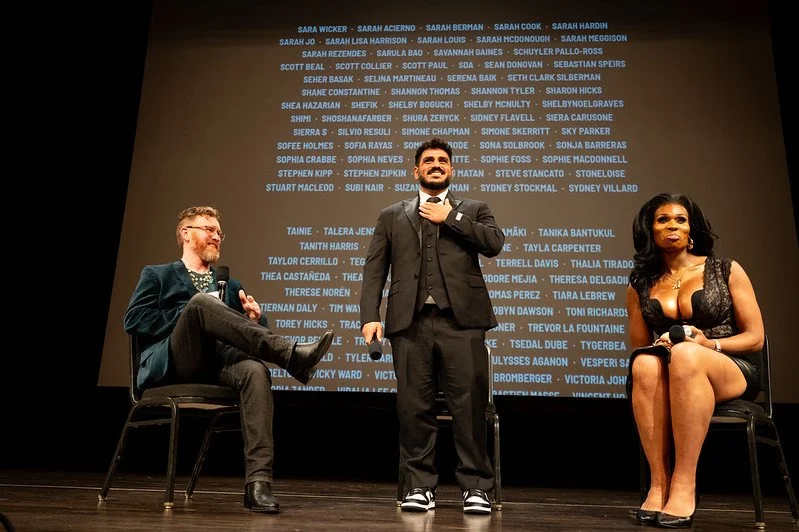 Three people on stage at an event, with a dark background filled with numerous names in blue text. Two are seated, and one is standing, holding a microphone and smiling.