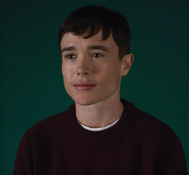 Portrait of a young woman with short dark hair and freckles, wearing a maroon sweater, against a green background.