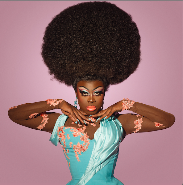 A woman with elaborate makeup and a large afro hairstyle, wearing a turquoise dress with pink lace details and matching arm accessories, posing against a pink background.