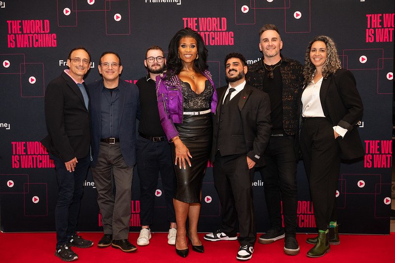 Group of seven people standing in front of a backdrop with the text "The World is Watching" and media icons, posing for a photo at an event.