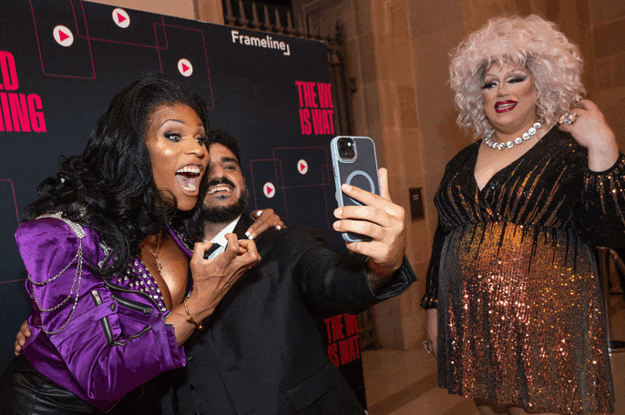 Three people taking a selfie at an event, with a backdrop that reads "The We is What" and "FrameLine." The person on the left is wearing a shiny purple outfit, the person in the middle is dressed in black, and the person on the right has curly gray hair and is wearing a sparkly black and gold dress with a pearl necklace.