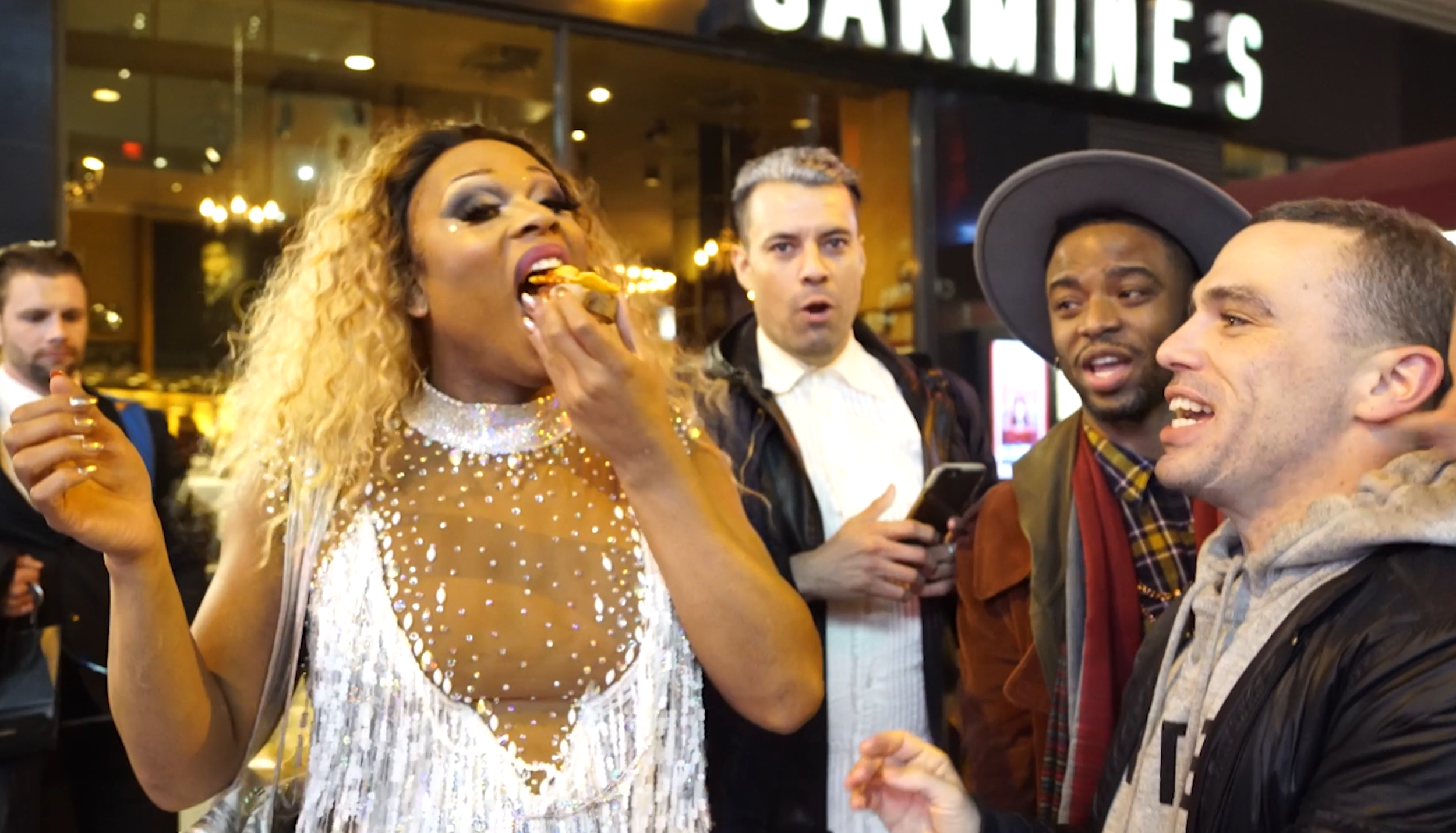 A group of people standing outside a restaurant or bar, with a woman in the center eating a slice of pizza and others watching her with surprised or amused expressions.