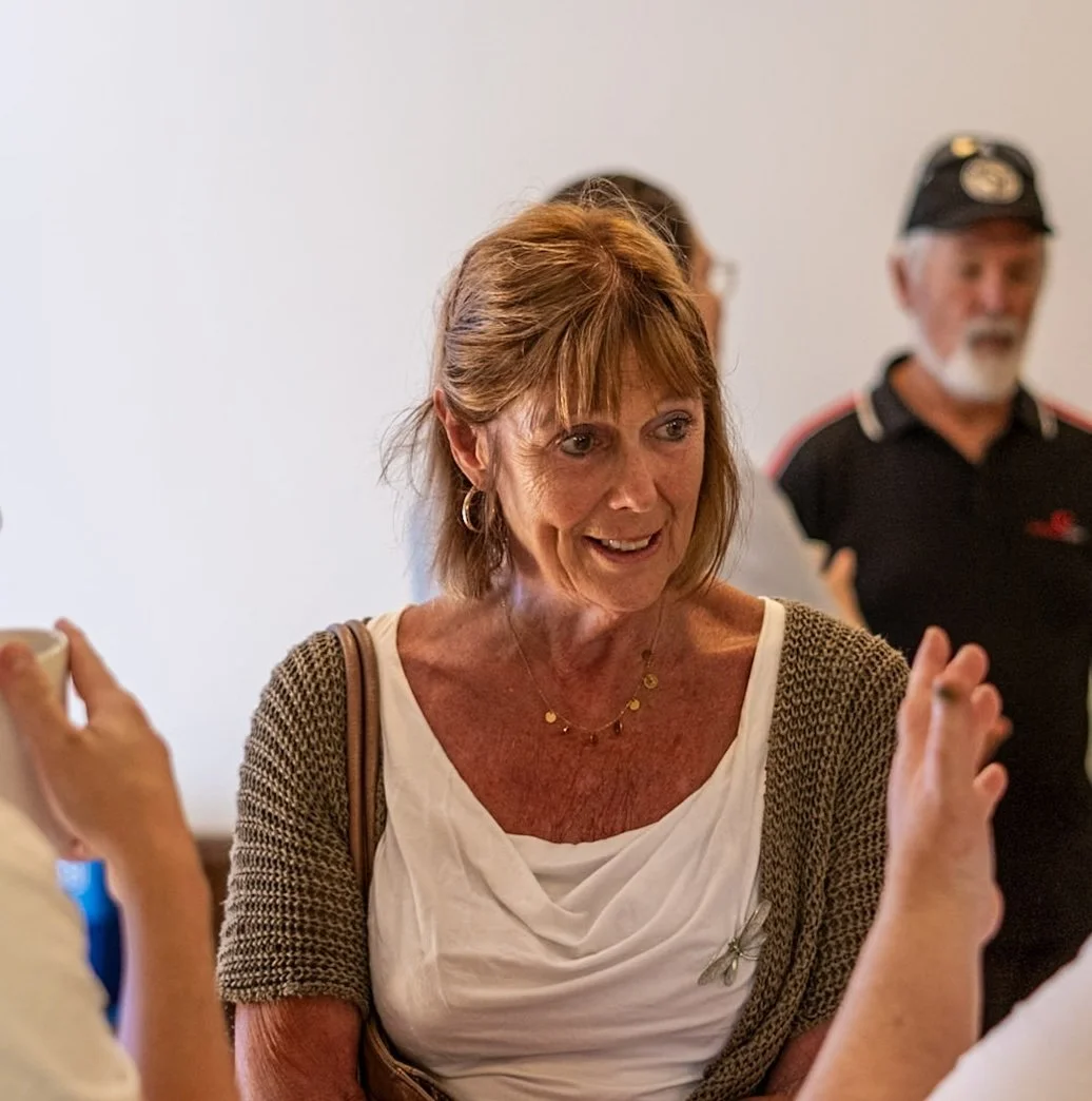 A woman with red hair smiling and talking with people at an indoor gathering, wearing a white top with a butterfly brooch, gold jewelry, and a brown cardigan, with a man in a black cap and shirt in the background.