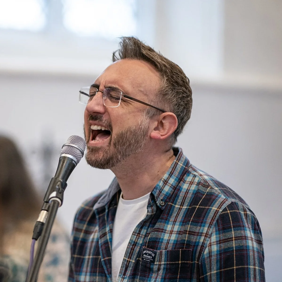 A man singing passionately into a microphone, wearing glasses and a plaid shirt, in an indoor setting.