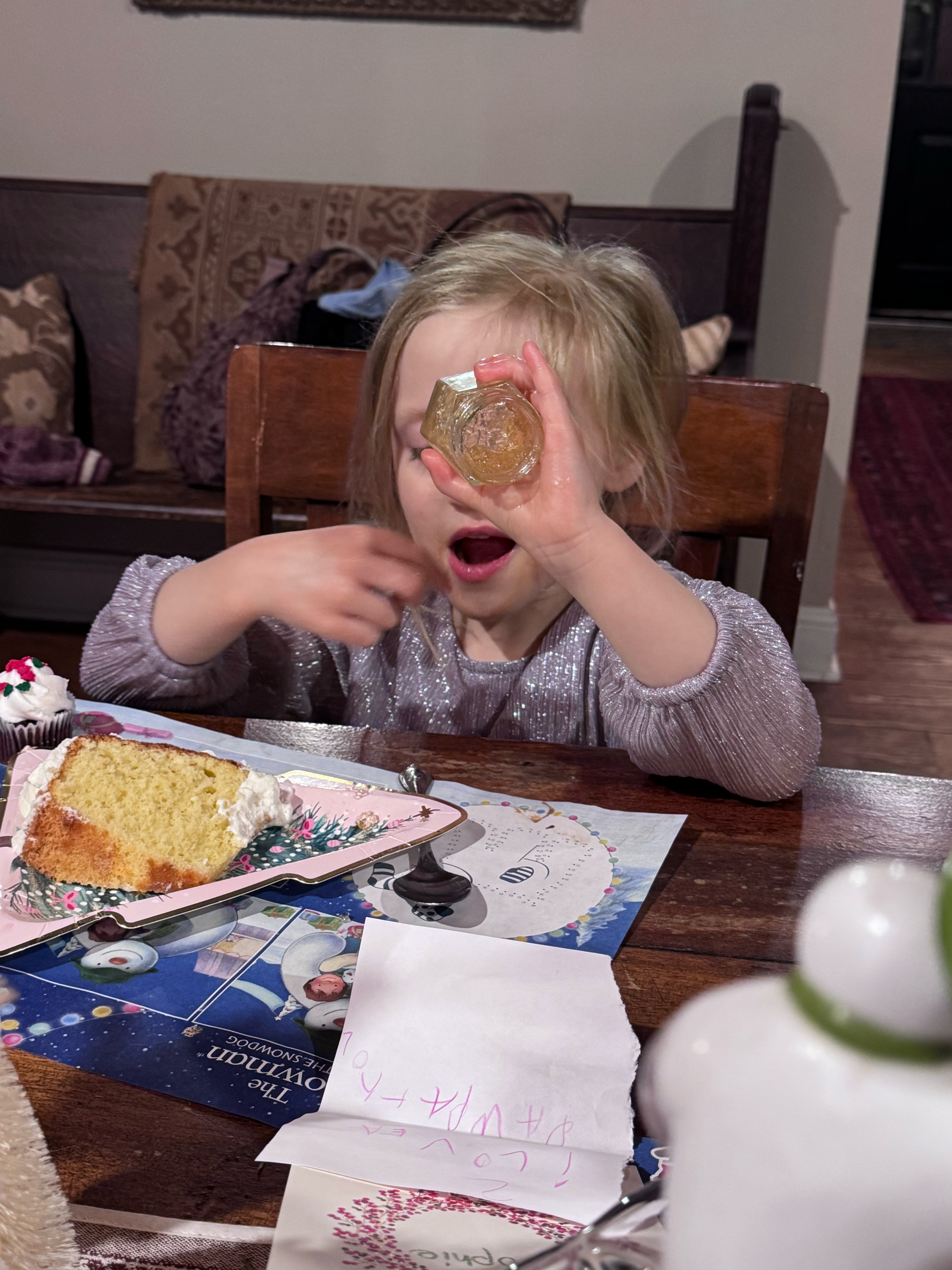 A young girl with blonde hair, wearing a sparkly silver top, sitting at a wooden table with a slice of cake and a cupcake. She holds an object up to her eye, looking through it with her mouth open. There are some papers and a colorful tablecloth on t