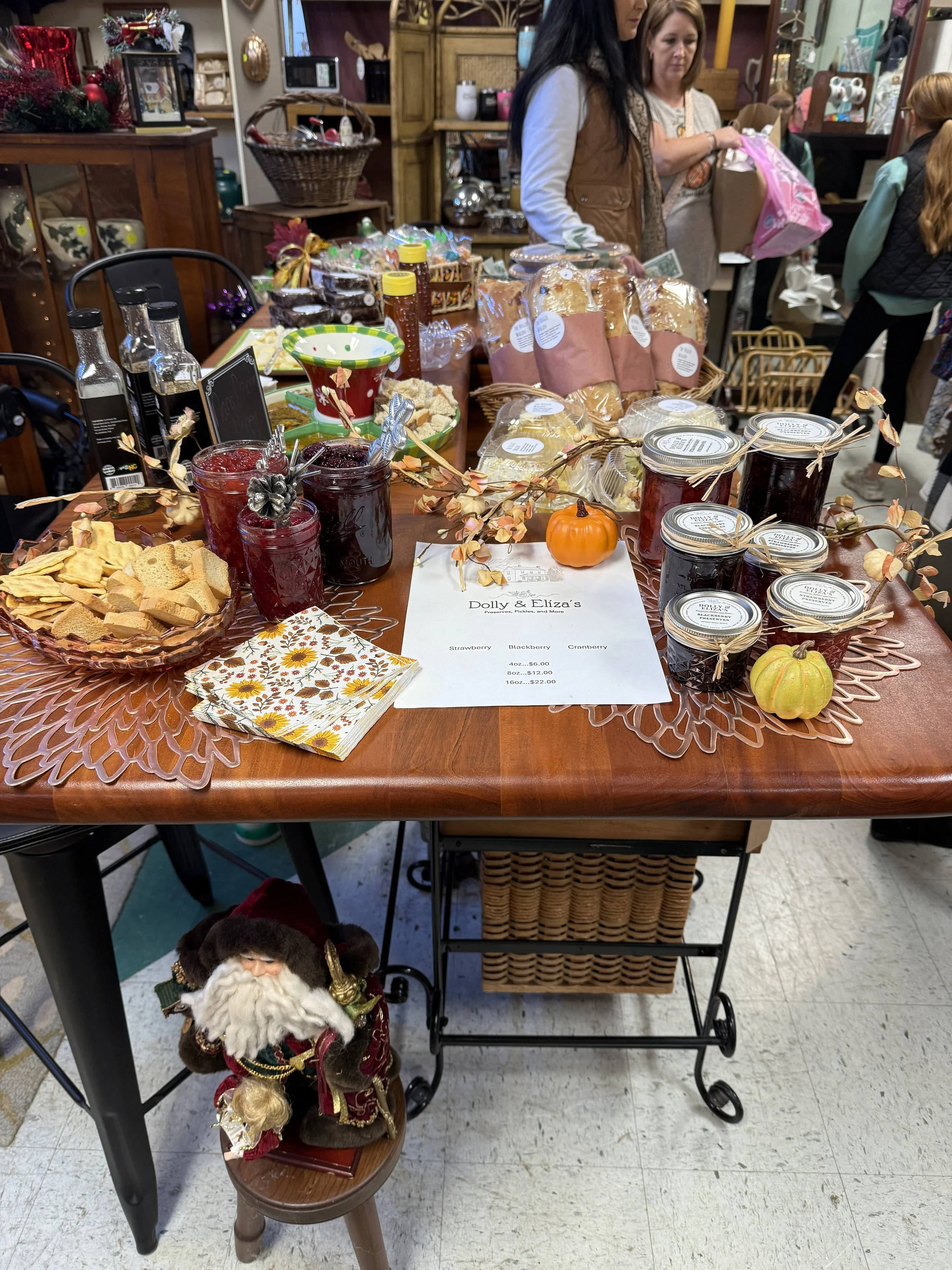 Table decorated with fall-themed items, including jars of jam, loaves of bread, and small pumpkins, at a holiday event.