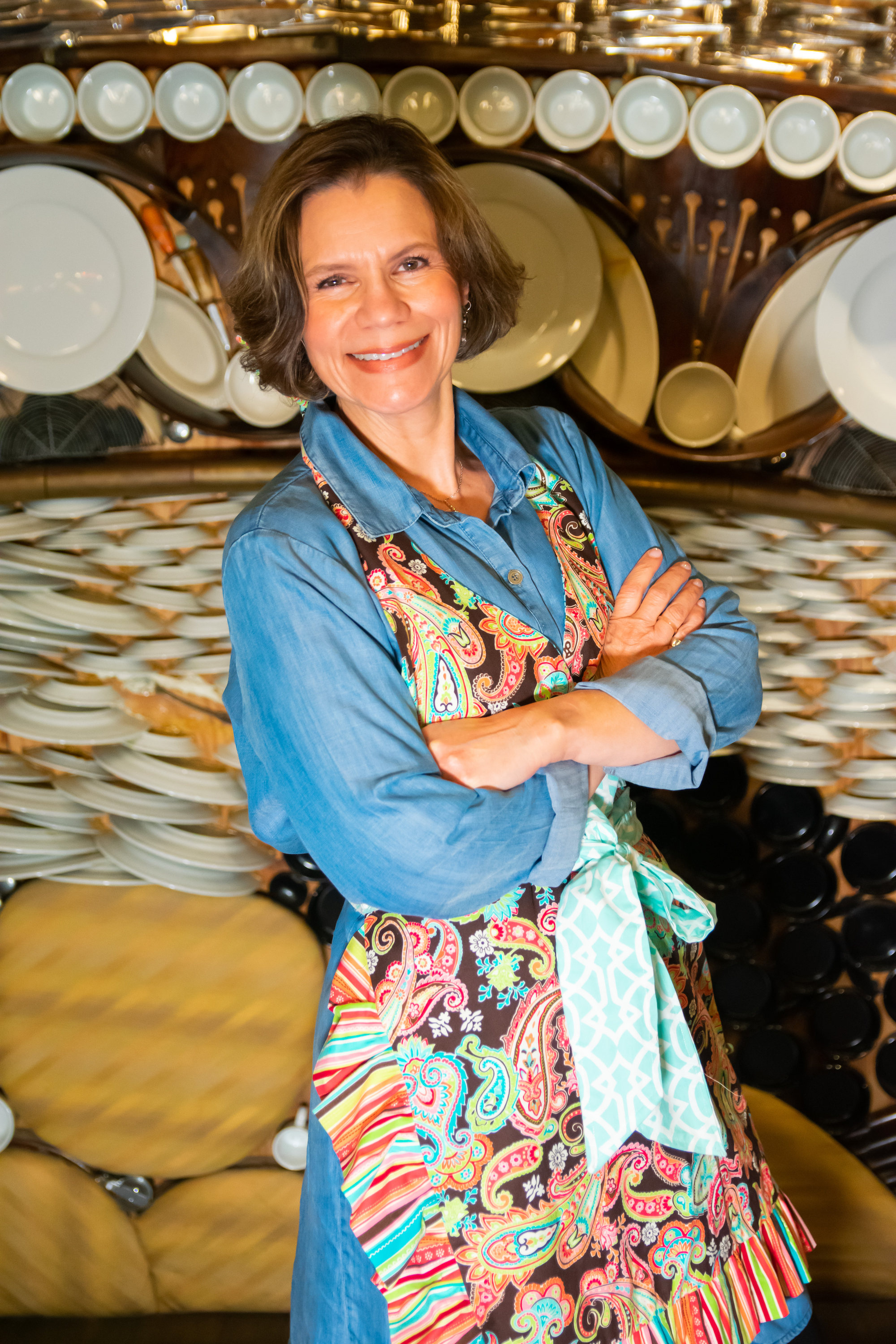 A woman in a blue shirt and colorful apron standing with arms crossed in front of a background of stacked dishes and plates.