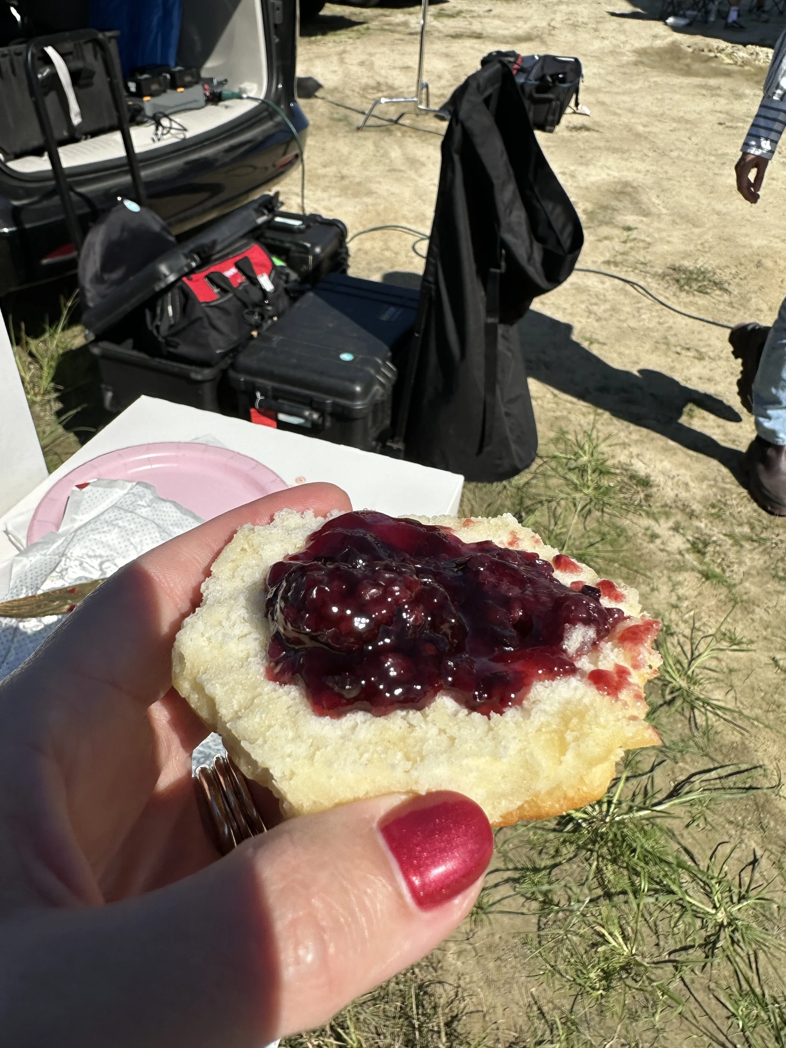 Hand holding a piece of bread with cream and berry jam on top, outdoors with equipment and luggage in the background.