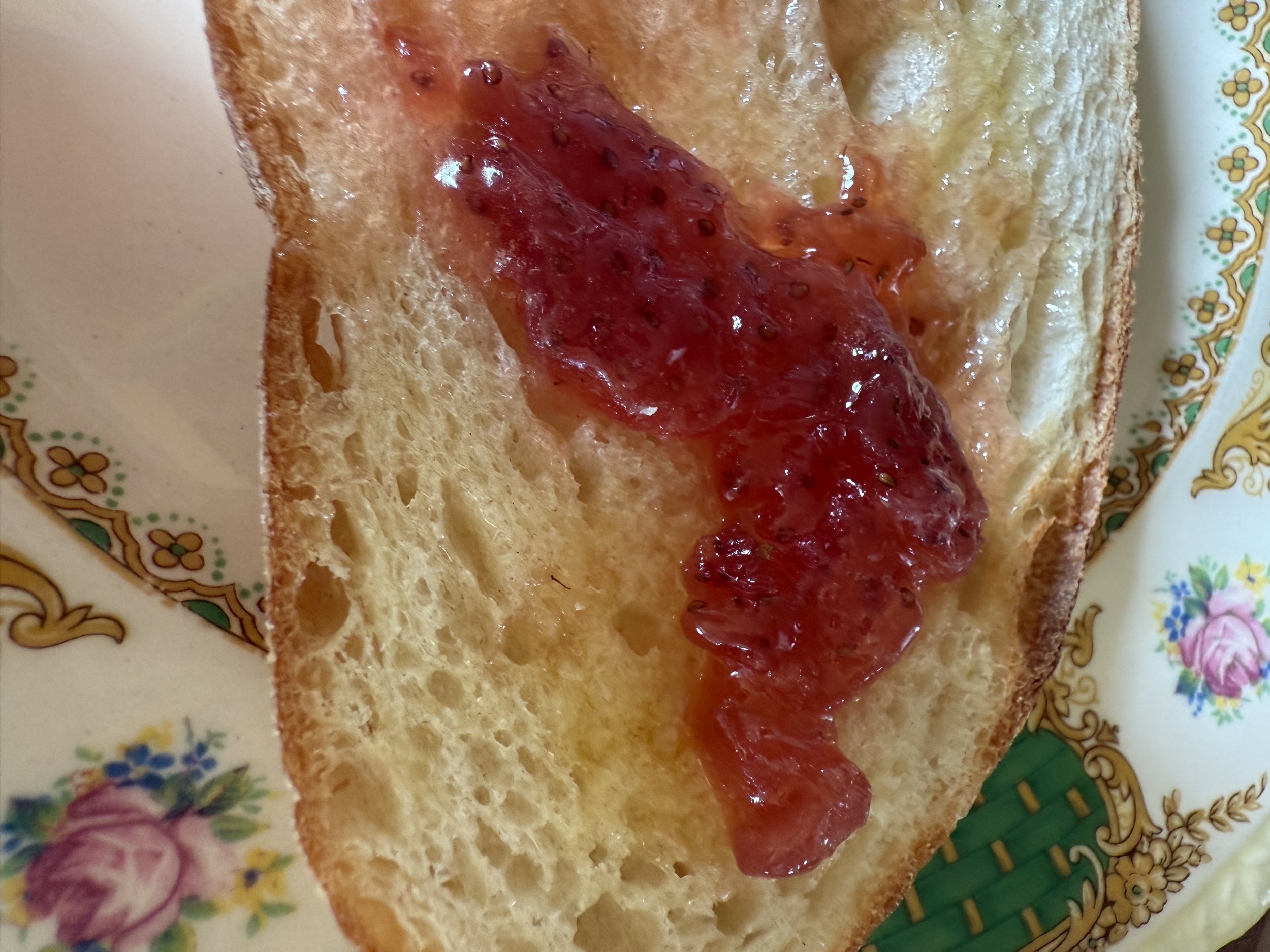 Close-up of a slice of toasted bread with red jam spread on top, placed on a decorative floral-patterned plate.