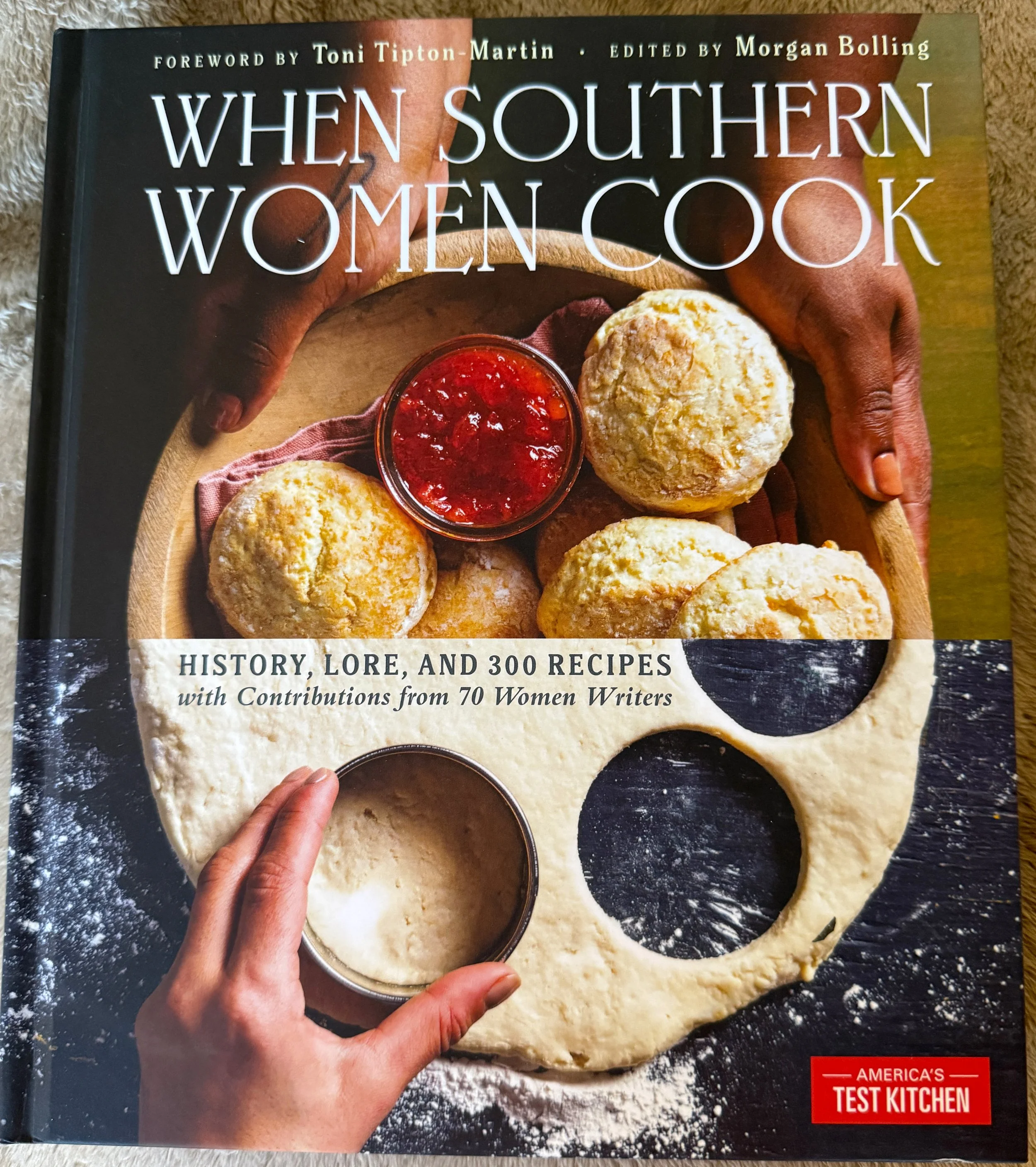 Cover of a cookbook titled 'When Southern Women Cook.' The cover features a wooden tray with biscuits, a small bowl of red jam, and a hand holding a biscuit with a biscuit dough circle on a black surface with flour. The book includes history, lore, 300 recipes, and contributions from 70 women writers, published by America's Test Kitchen.