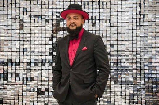 Male attorney in black suit with red shirt and matching hat standing in front of a reflective mosaic wall.