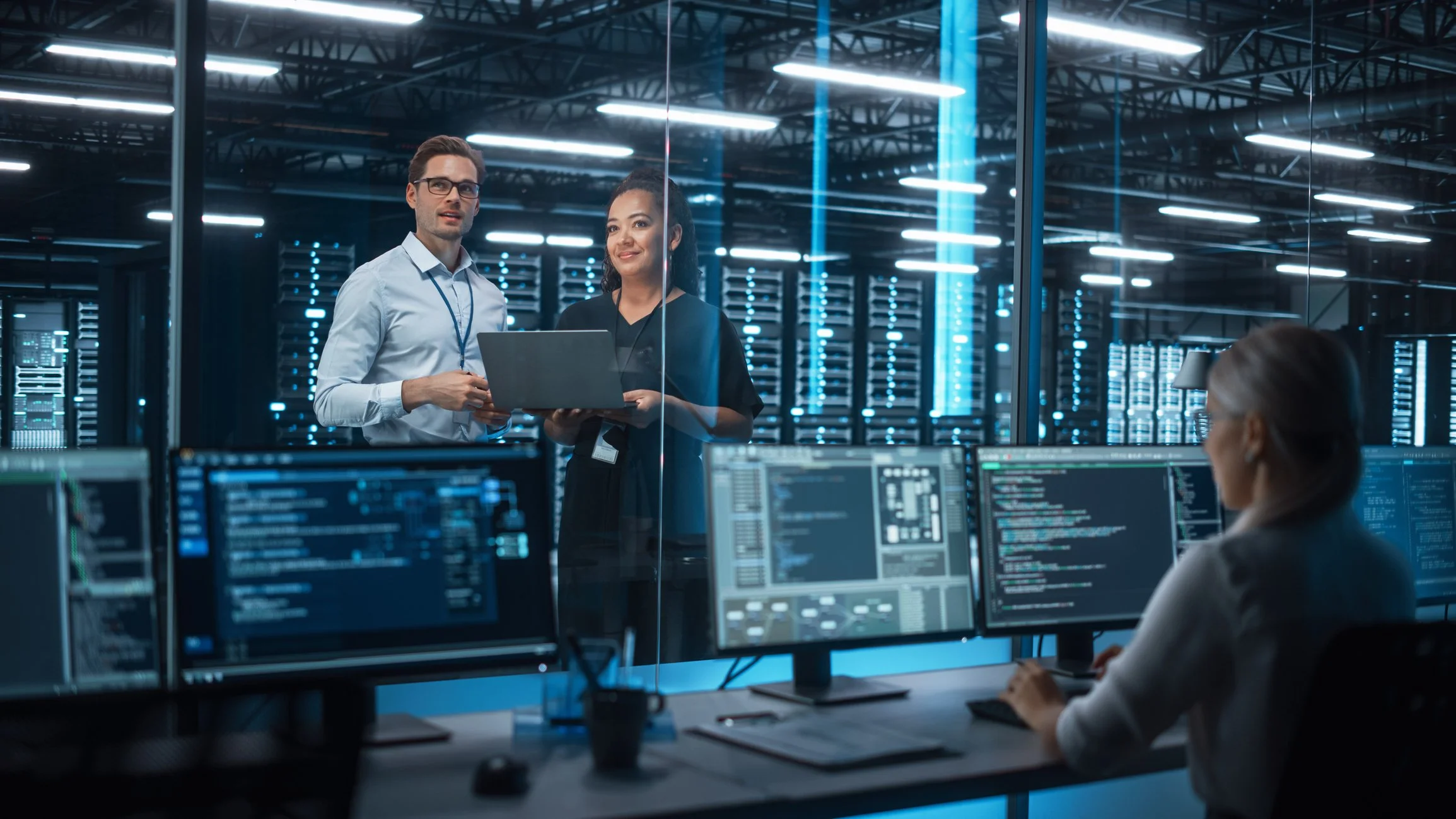 Team of three IT professionals working in a server room with multiple computer monitors displaying code and data.