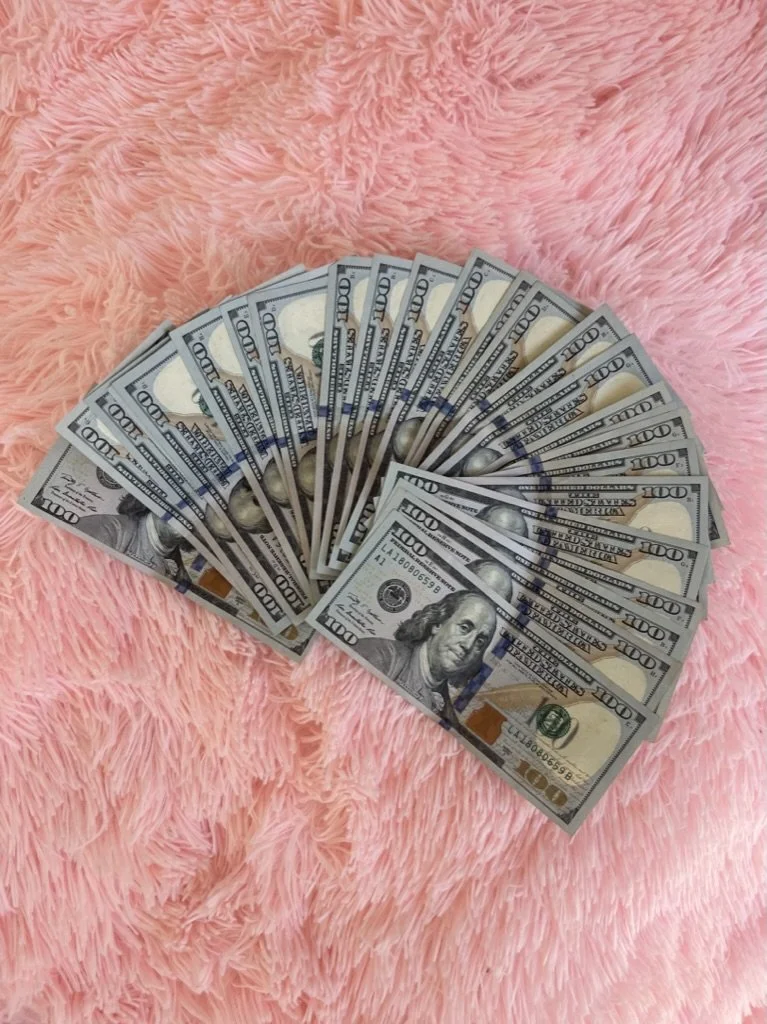 Fanned out hundred dollar bills held laying on a pink fluffy blanket.