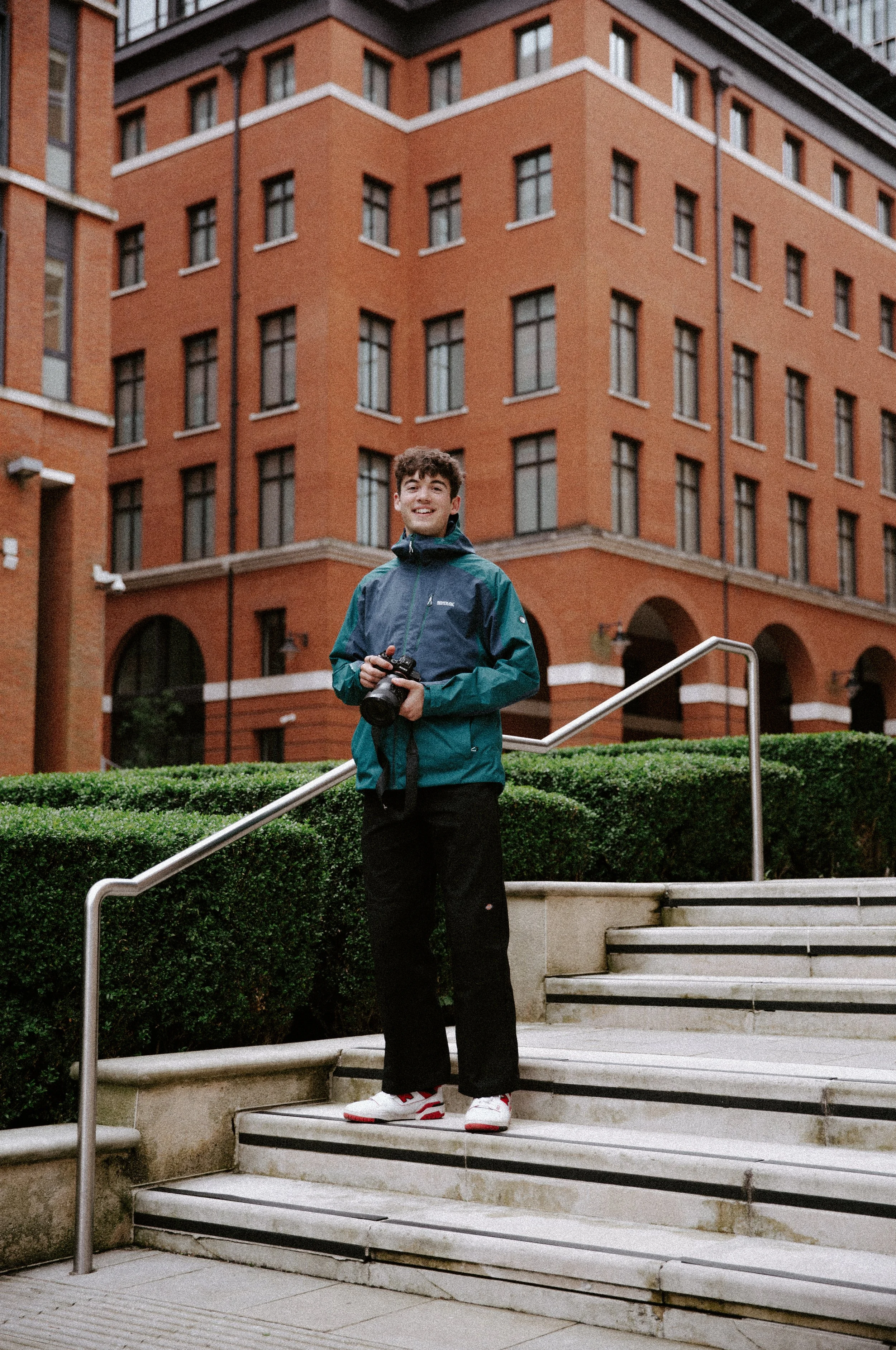 A young man standing on outdoor stairs holding a camera, with a red brick building and greenery in the background.