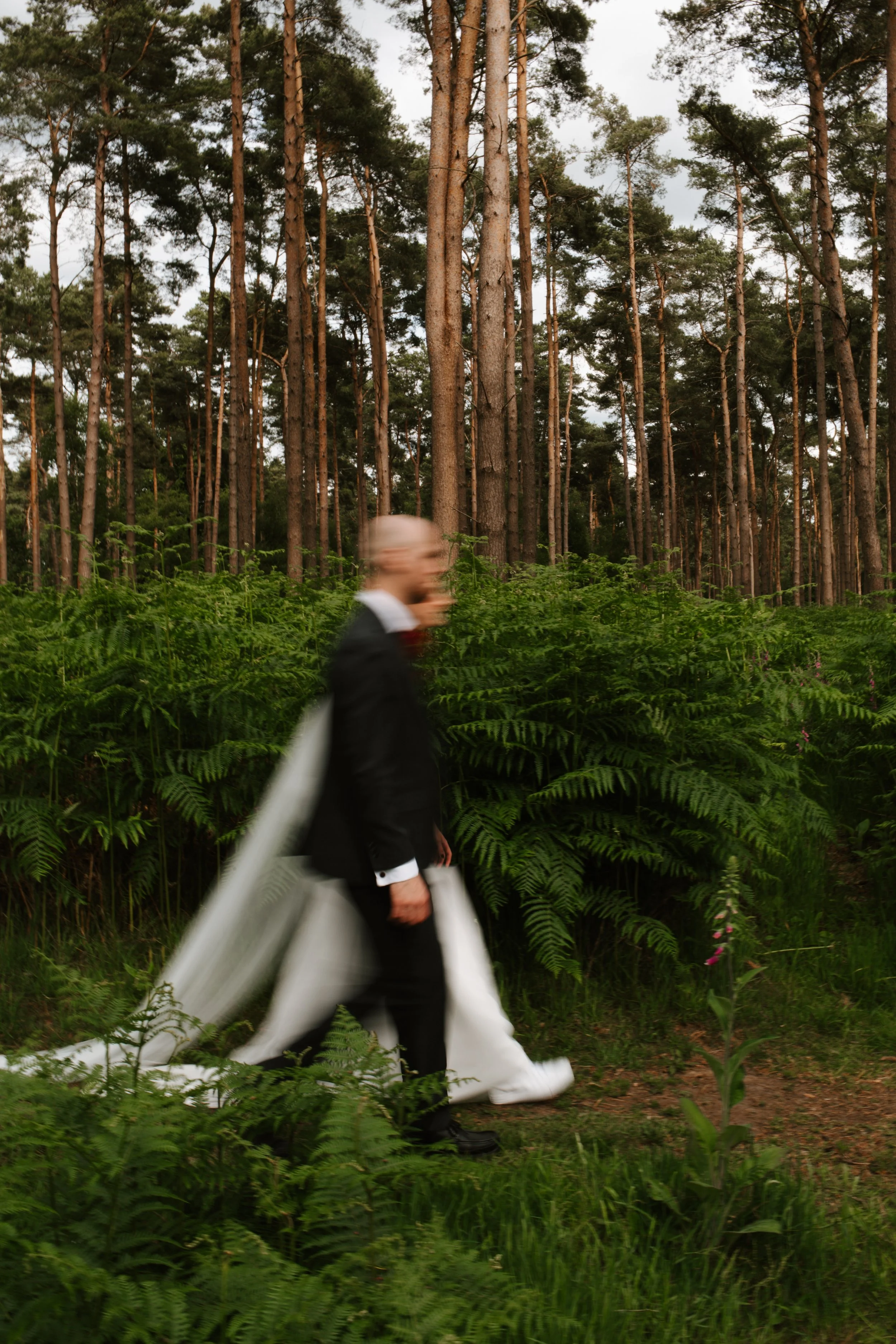 A person dressed in a black suit with white shoes and a white cape is walking through a forest with tall trees and green foliage.