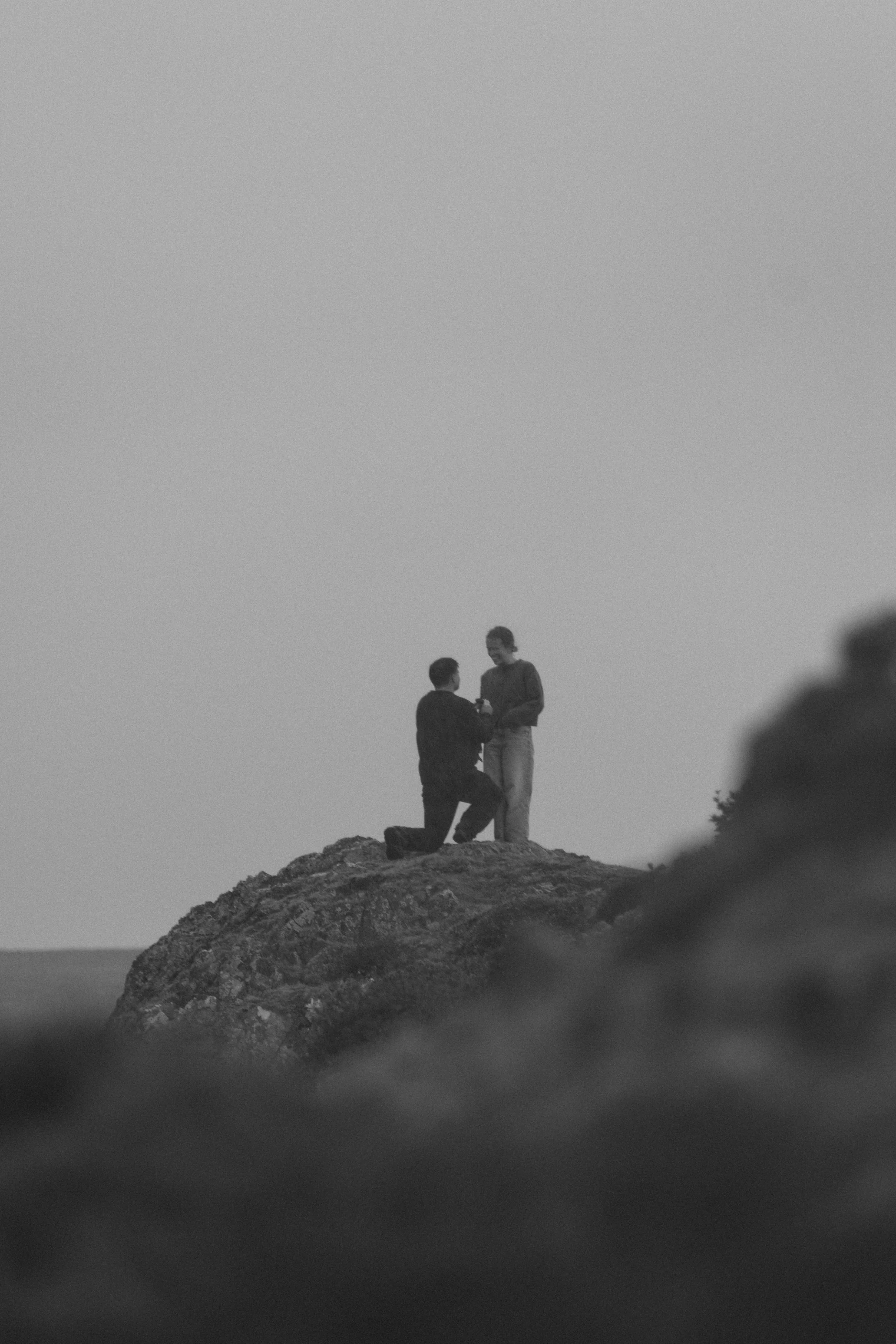 Two men on a rocky hilltop, one kneeling and proposing marriage to the other.