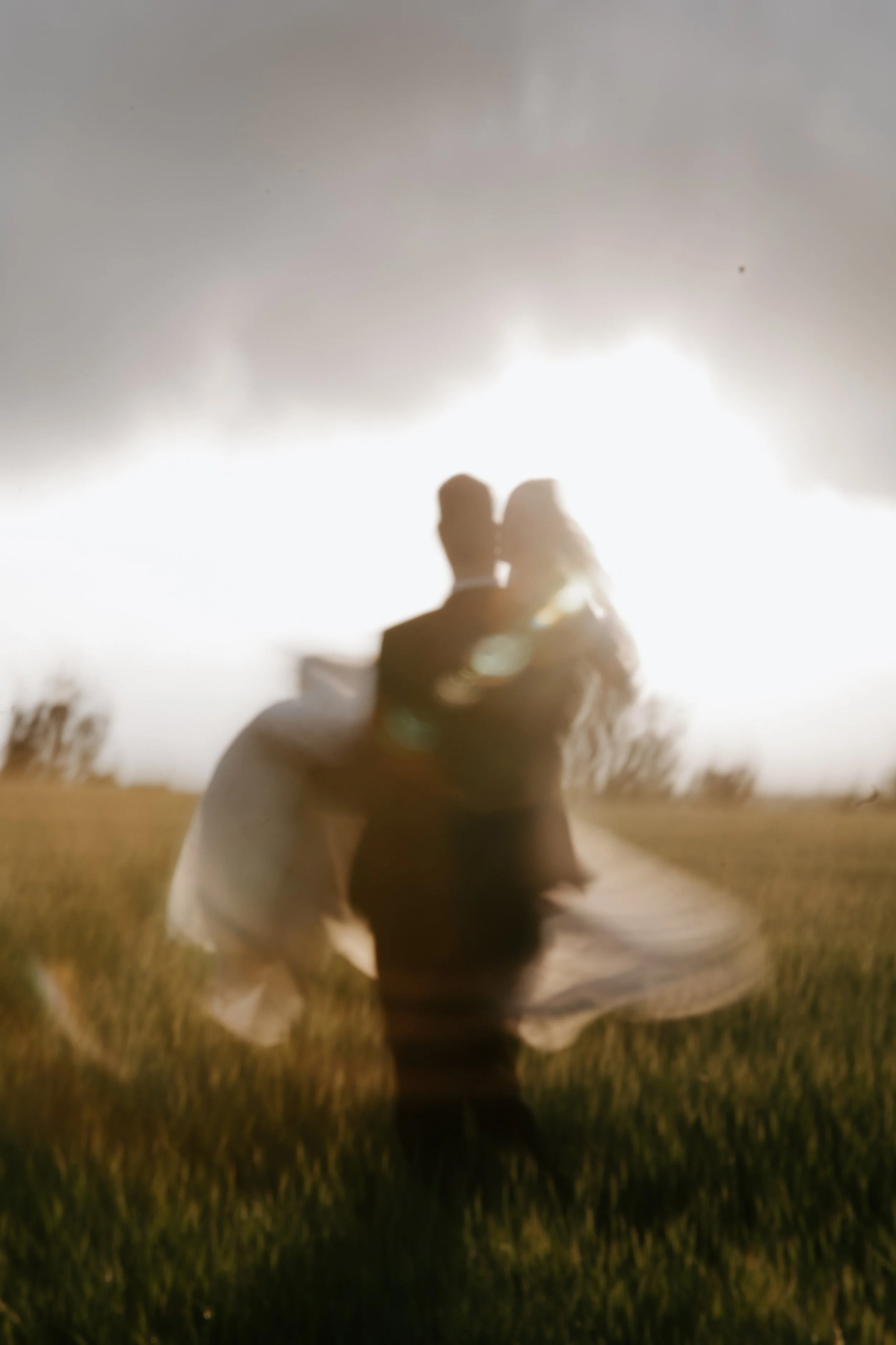 Silhouetted couple hugging in a grassy field with the sun setting or rising behind them, creating a bright, backlit scene with a romantic and dreamy atmosphere.
