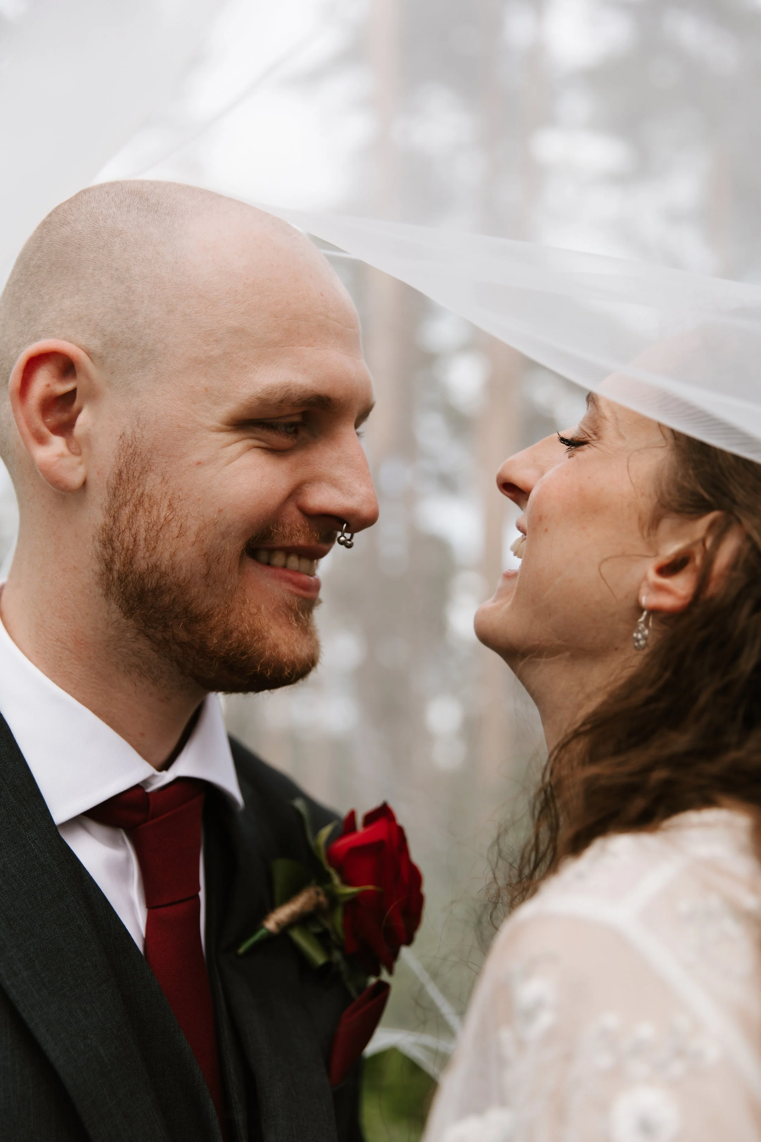 A smiling couple on their wedding day, close together outdoors, with the bride wearing a veil and earrings, and the groom in a suit with a red tie and boutonniere.
