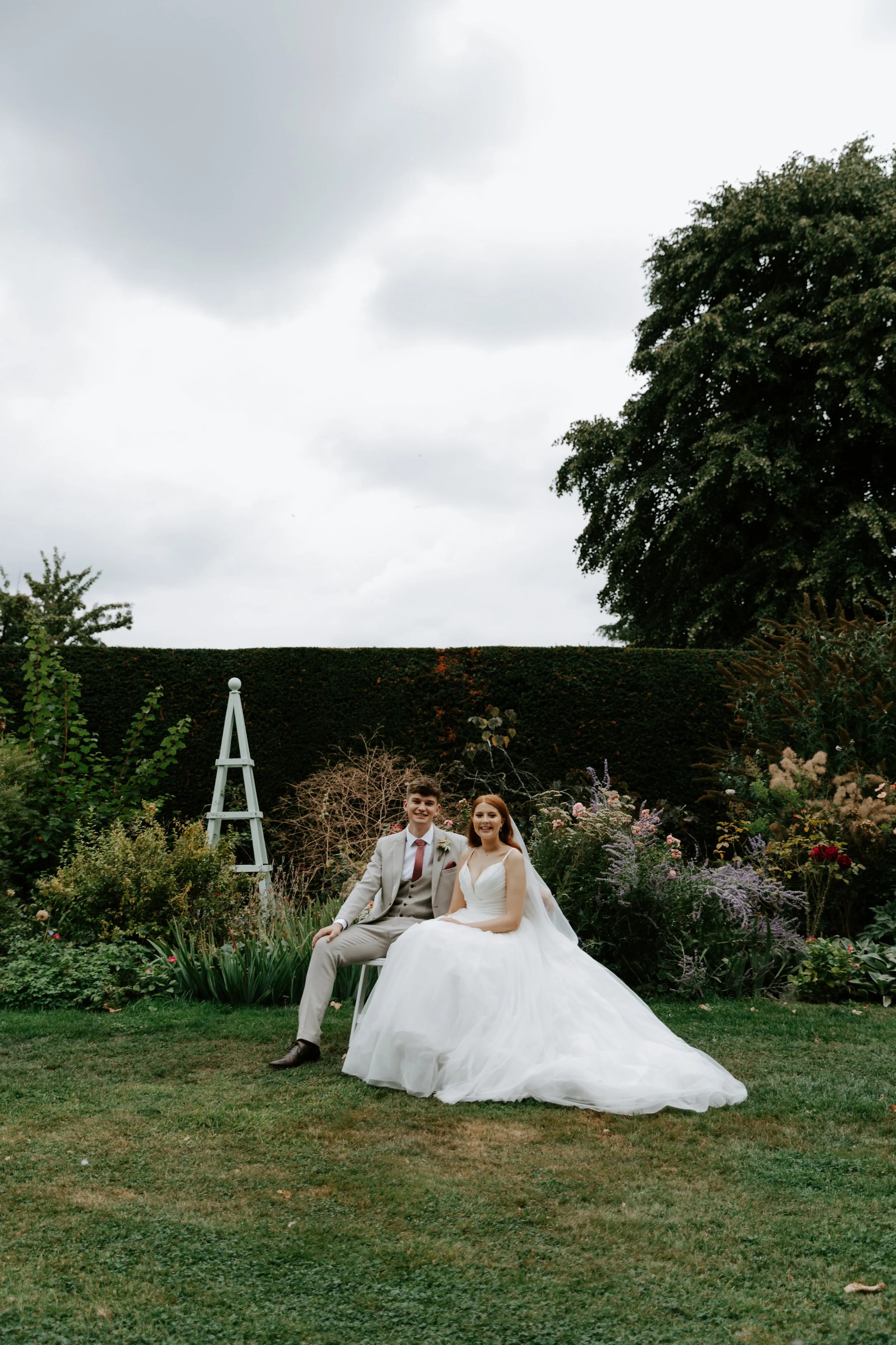 Bride and groom sitting in a garden with greenery and flowers, outdoor wedding photo, overcast sky.