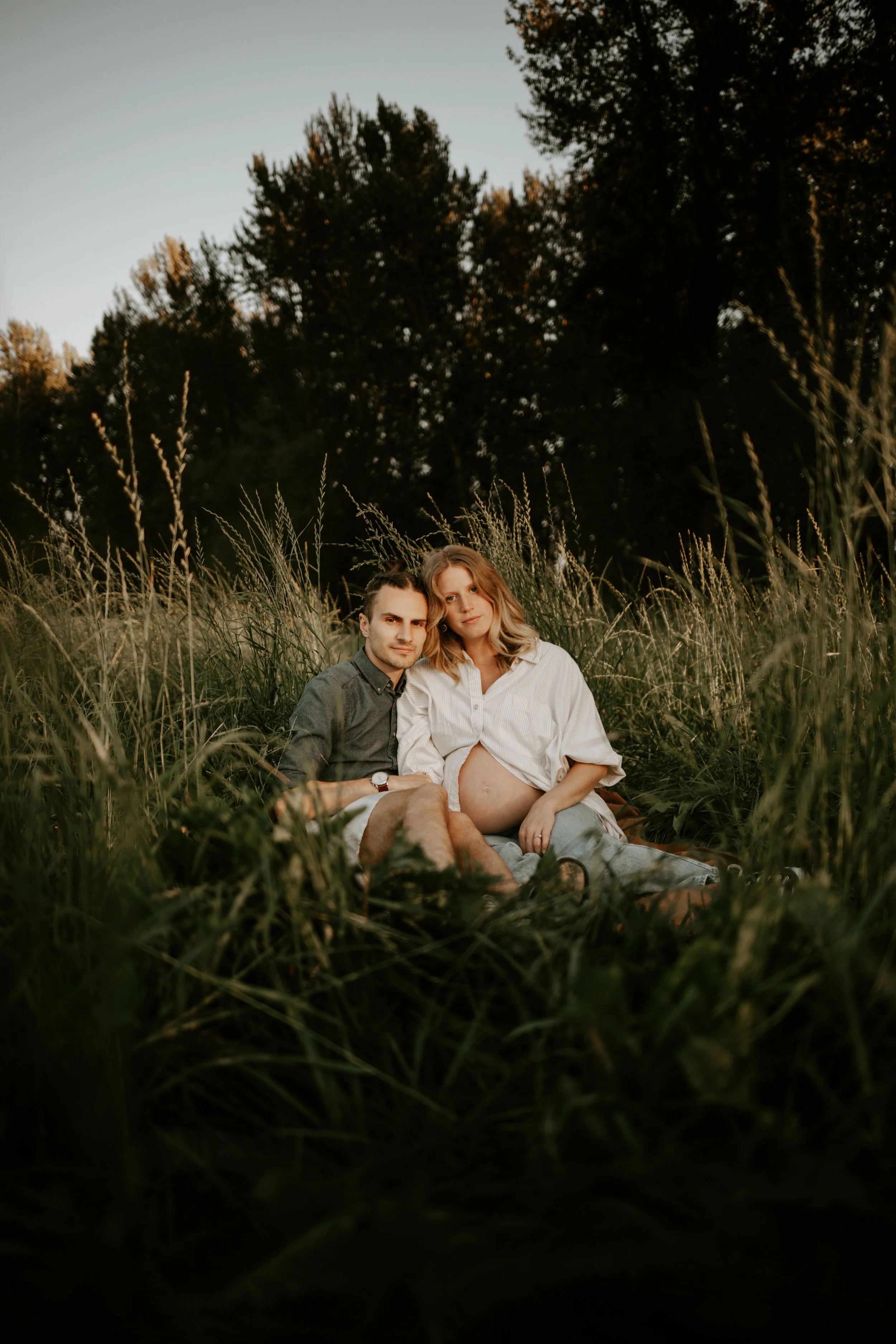 A pregnant woman and a man sitting in a grassy field during sunset, close to each other, with trees in the background.