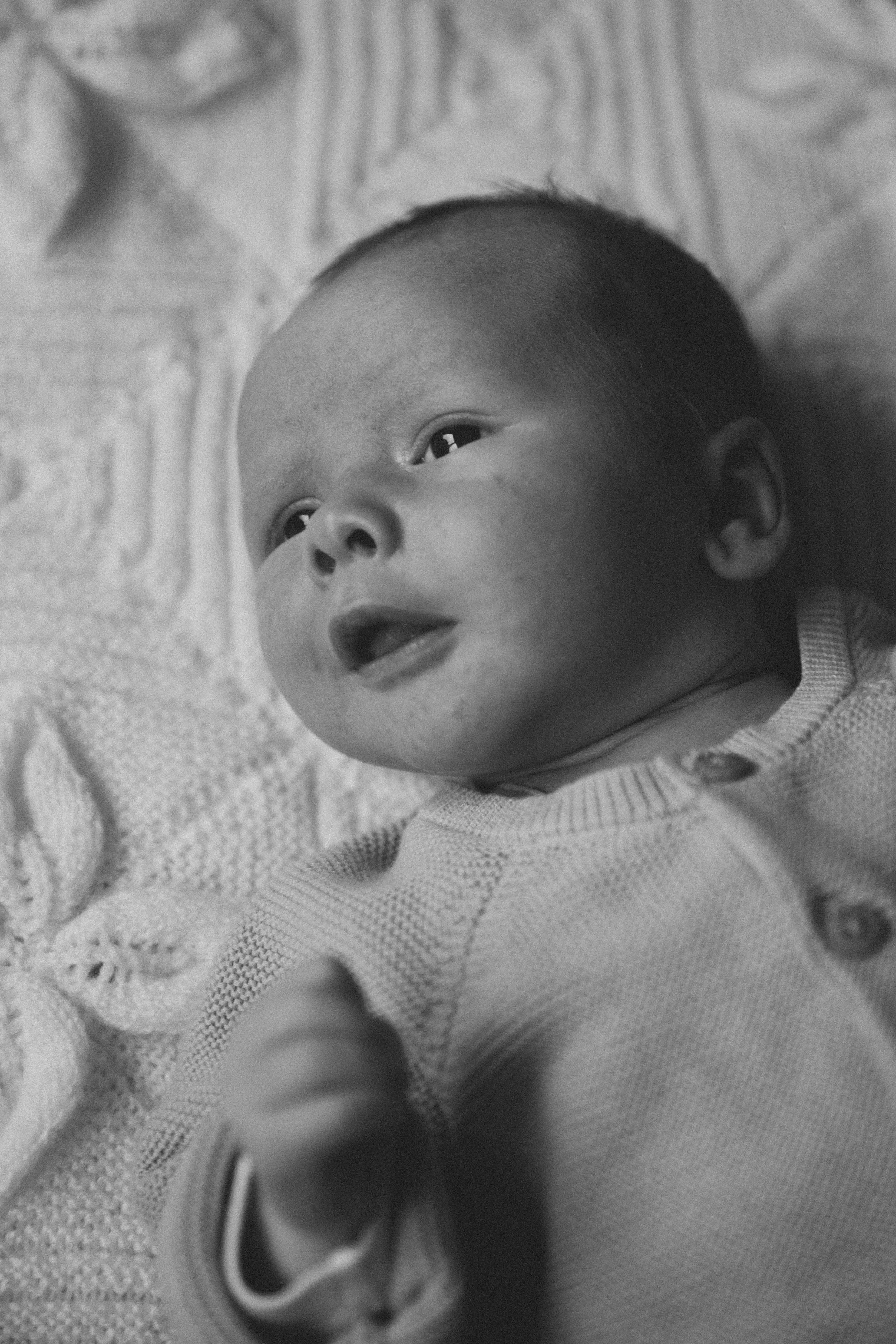 A black-and-white photo of a baby lying on a textured blanket, looking upward with a curious expression.