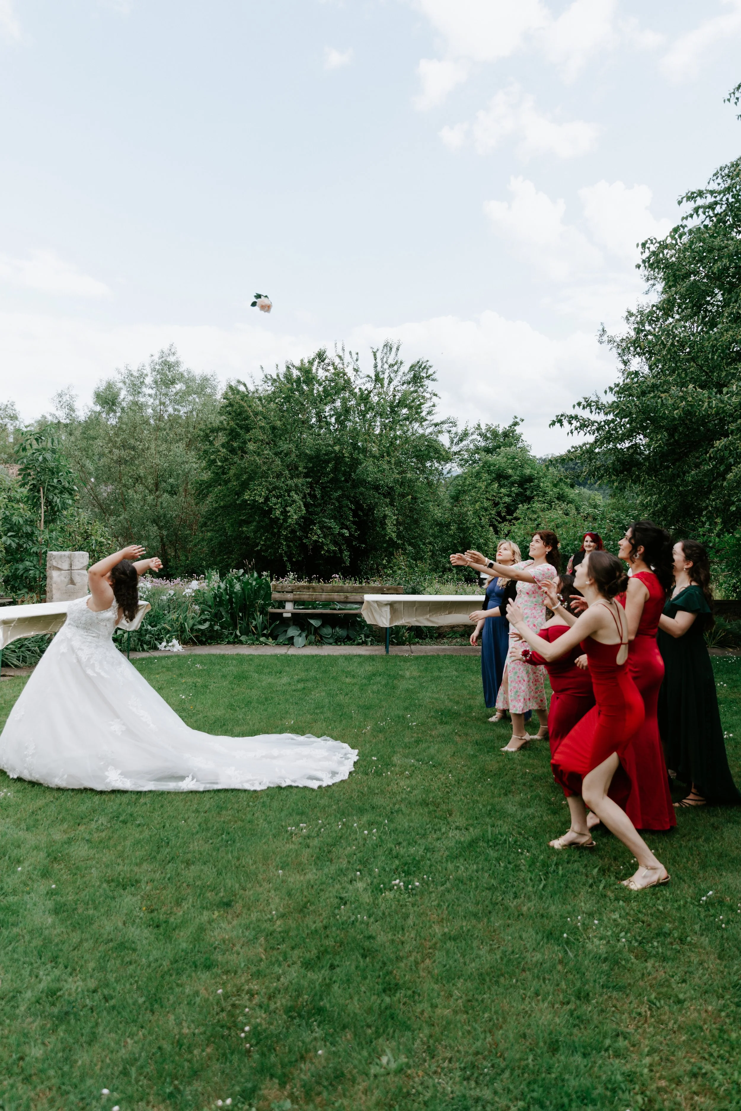 A bride in a white wedding dress throws a bouquet towards a group of women in colorful dresses, outdoors on a grassy area.