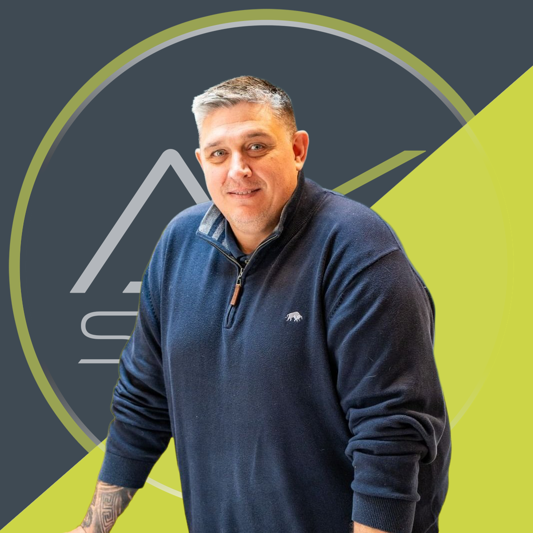 A man with short gray hair wearing a dark blue pullover with a logo of a bear, sitting in front of a stylized graphic background with mountain and speedometer elements in green, gray, and black.