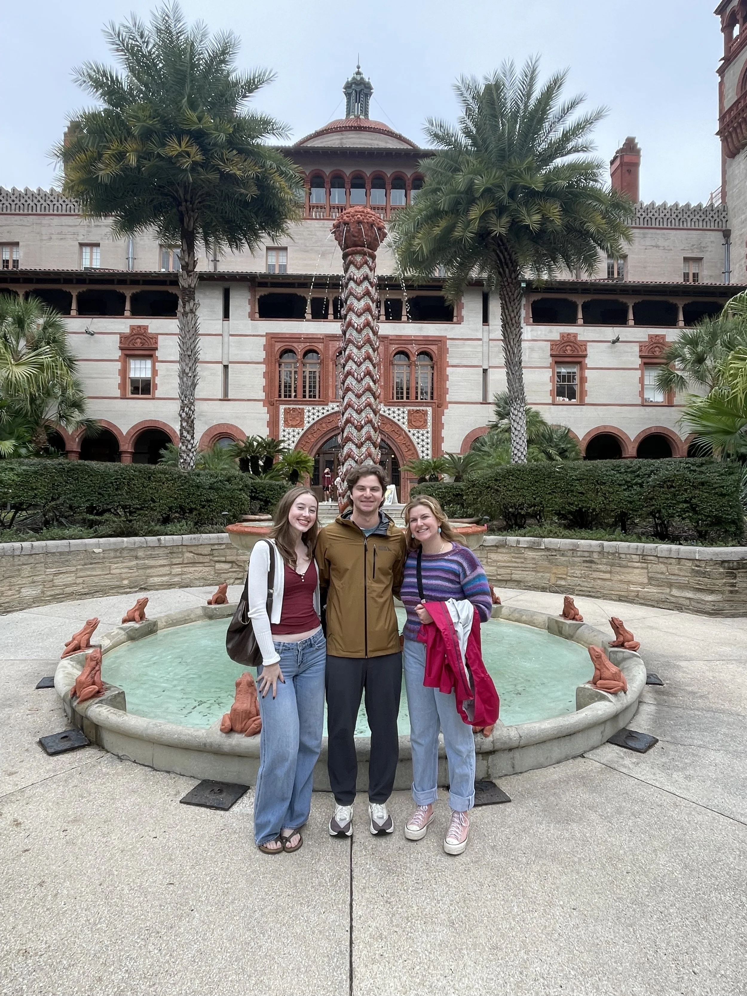 Three people, two women and one man, standing together in front of a fountain with frogs around its edge, in front of a historic building with palm trees and a decorative palm-shaped fountain feature in the background.
