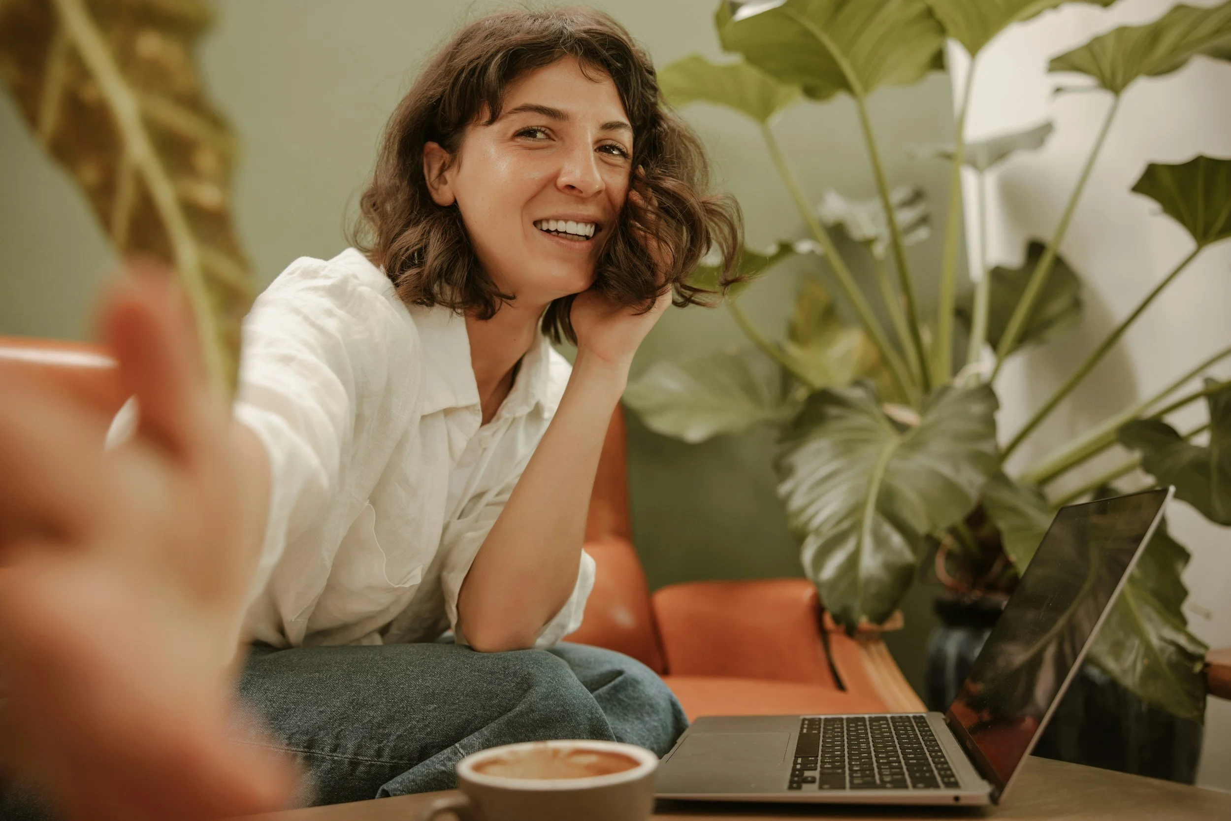 person with shoulder length brown hair, in front of laptop, plants in background