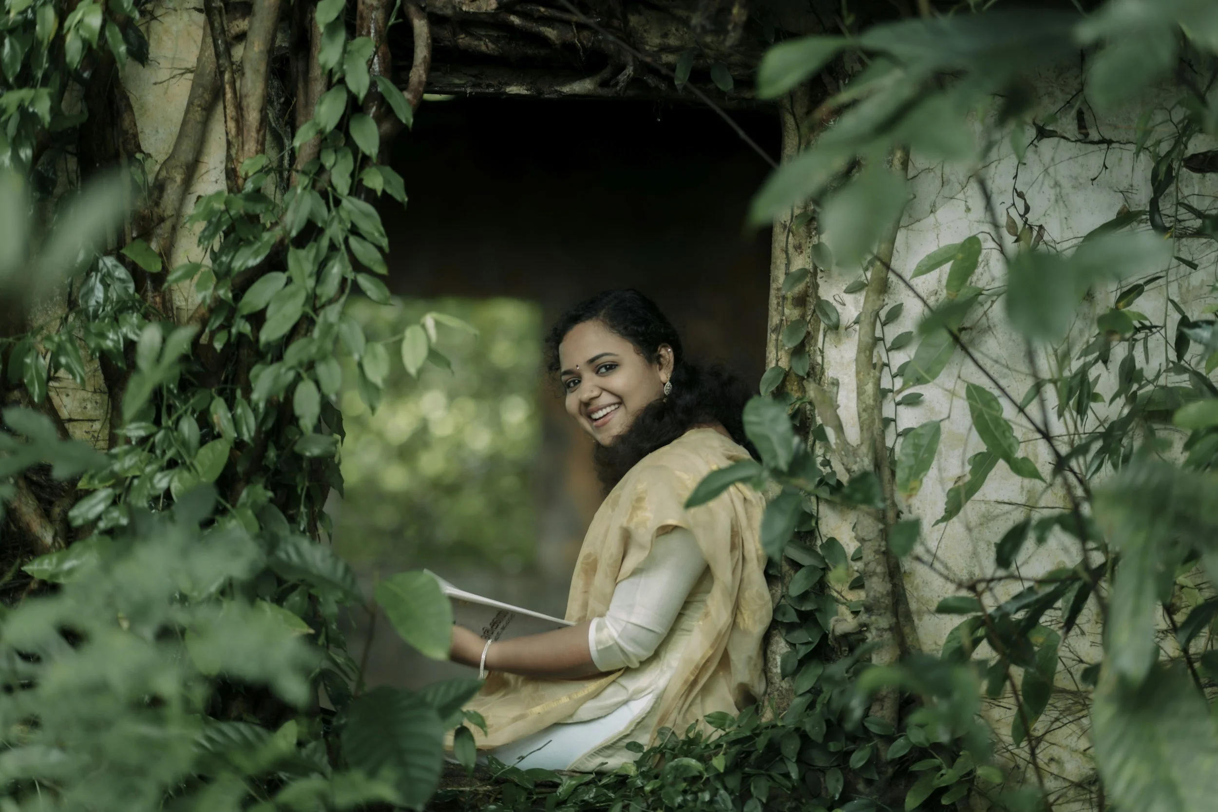BIPOC woman smiling, wearing yellow sari, sitting on bench in forest