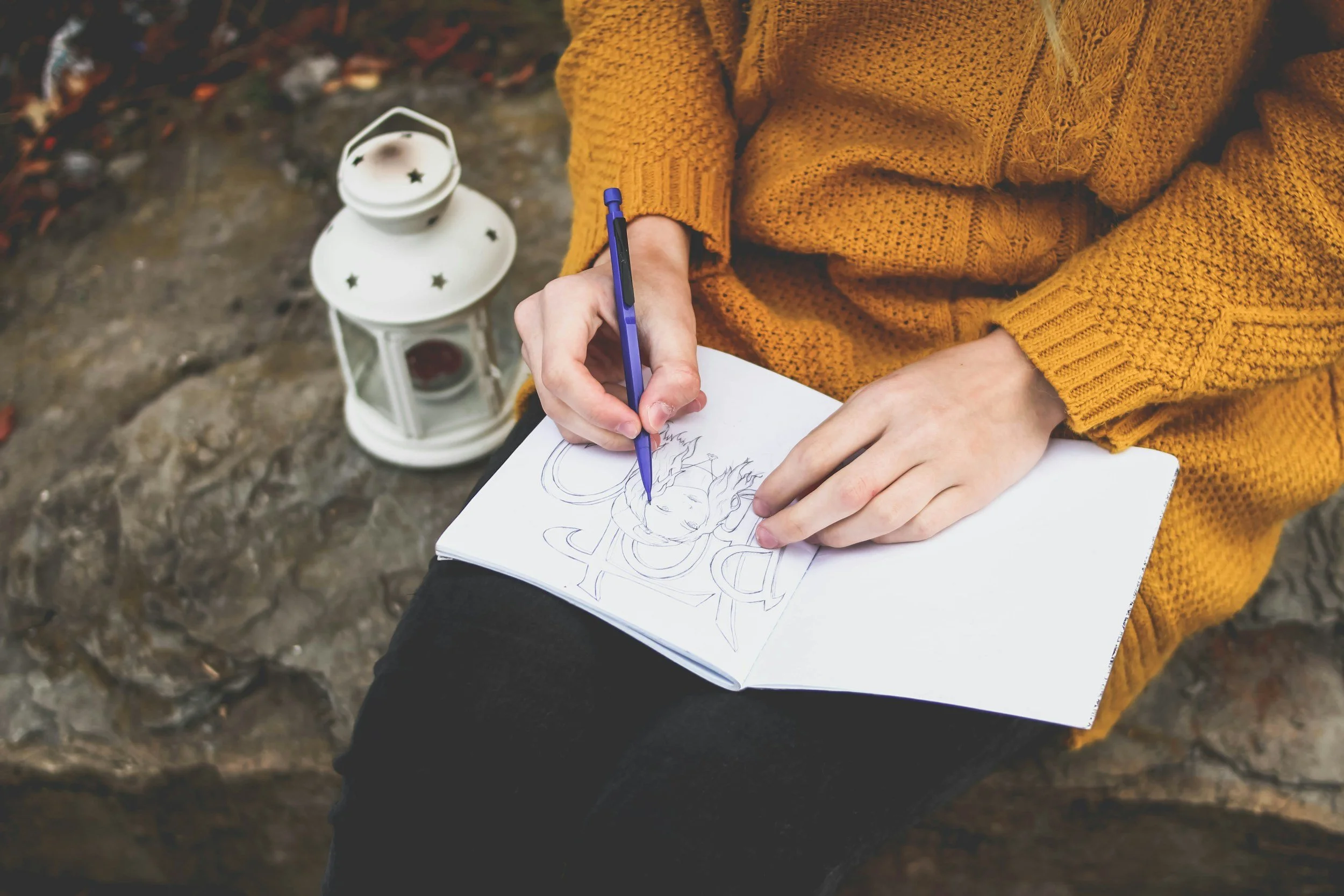 hands sketching image in journal, person wearing yellow sweater & sitting on rock
