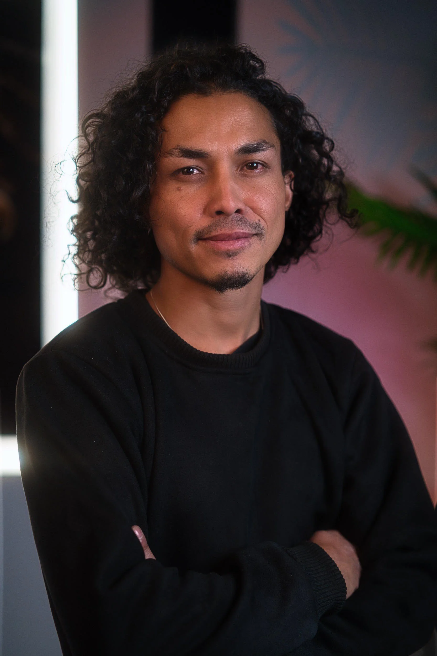 A man with curly dark hair, wearing a black shirt, standing with crossed arms in front of a colorful background with a plant in view.