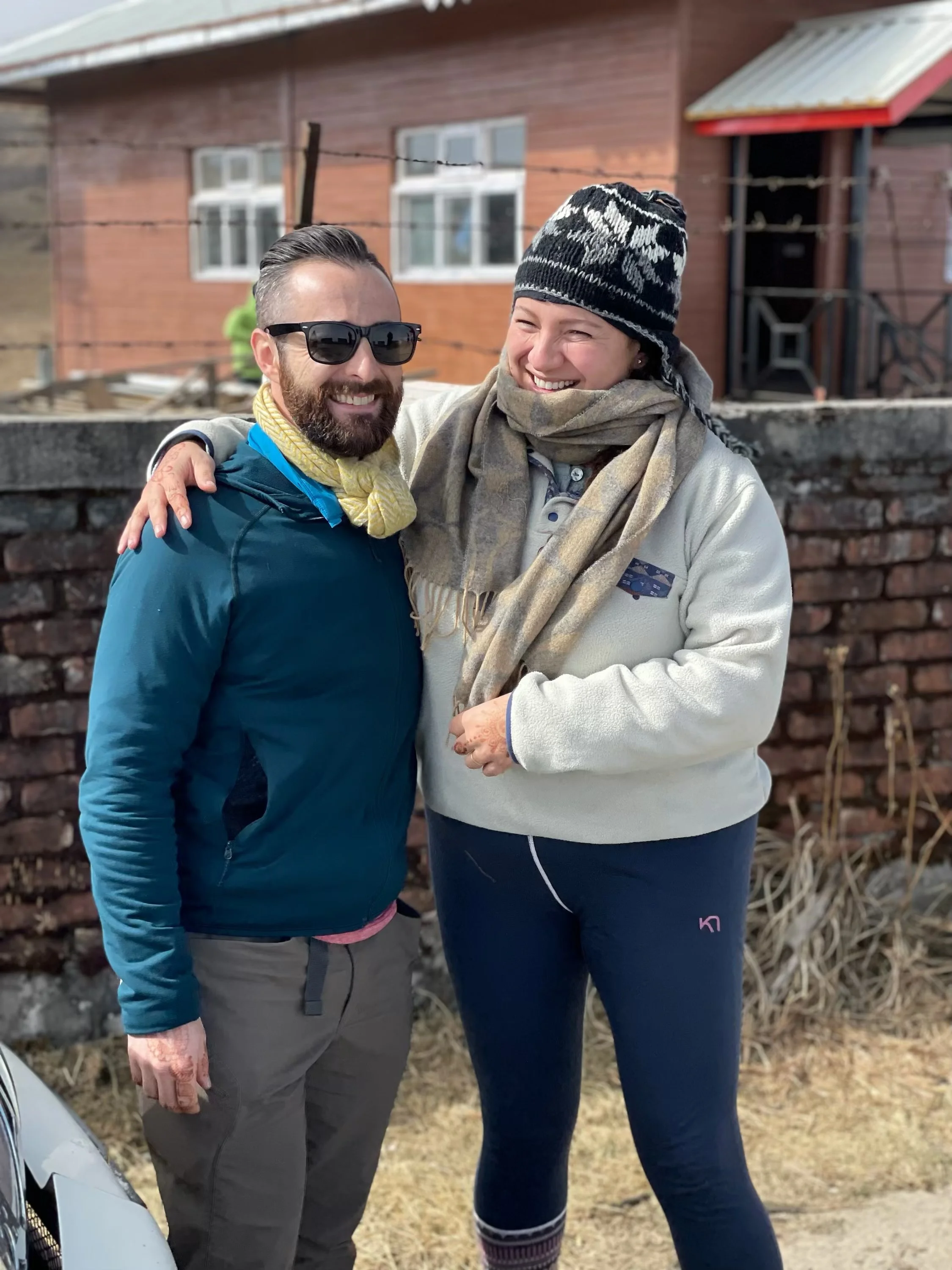 Two people, a man and a woman, smiling and embracing outdoors.