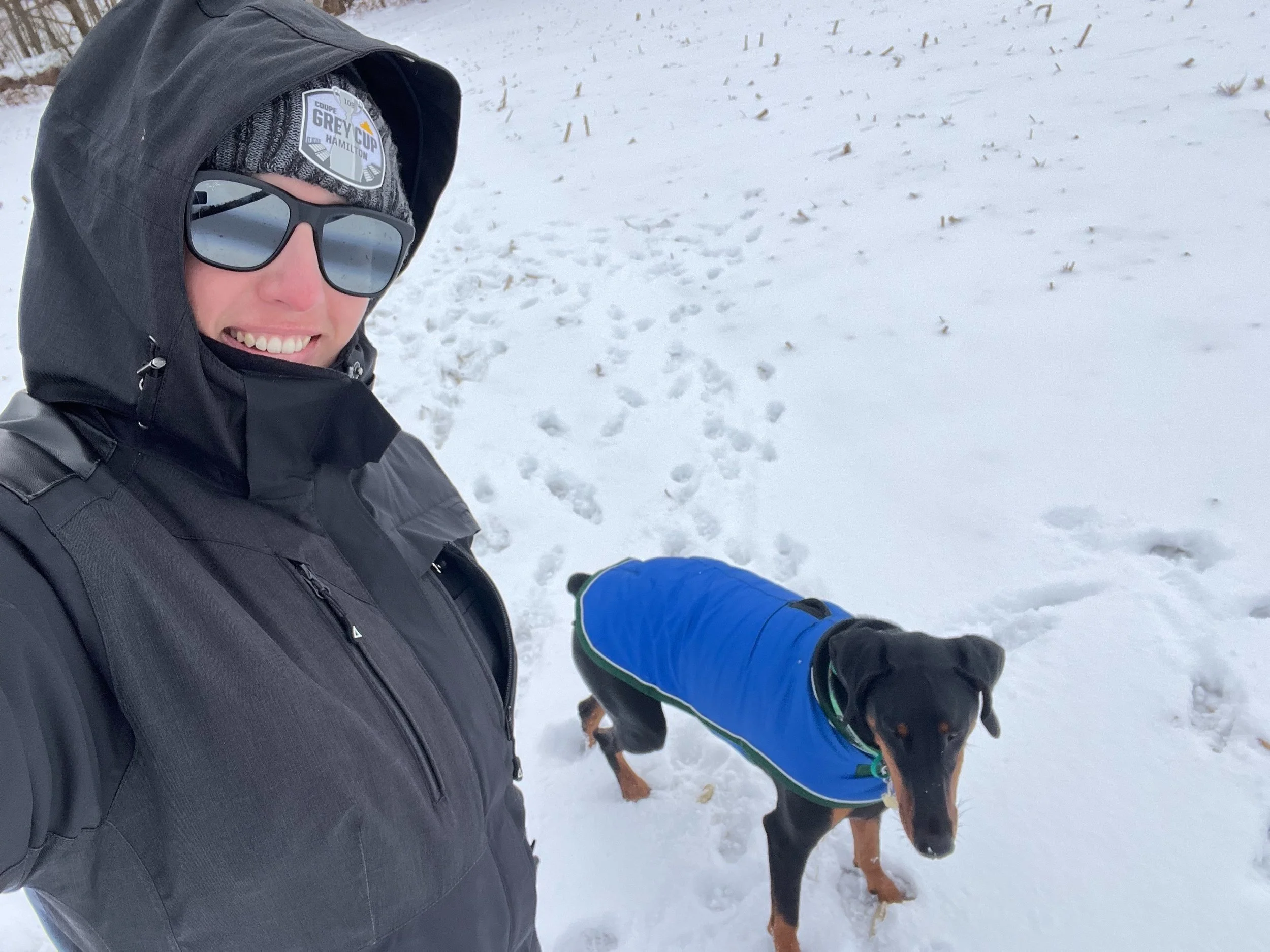 A person wearing a black jacket, sunglasses, and a grey beanie with a Patch labeled 'Grey Cup Hamilton' taking a selfie in a snowy outdoor area. A dog wearing a blue coat stands nearby.