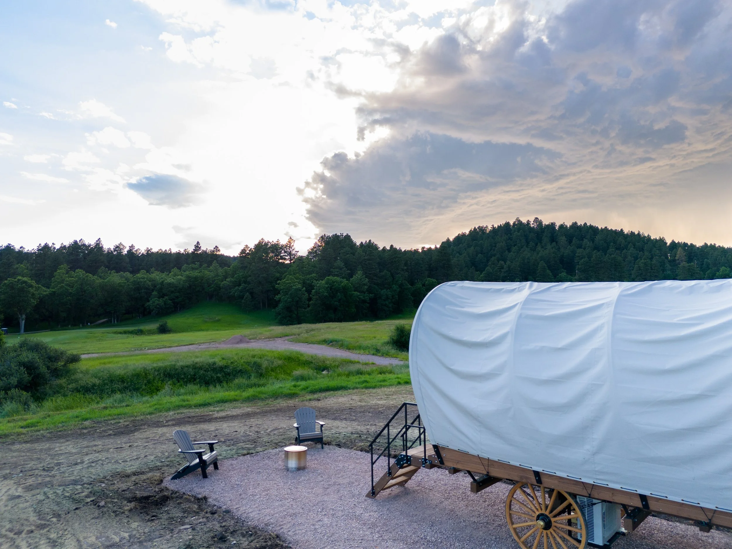 A covered wagon on a gravel pad with two Adirondack chairs and a small table, overlooking green fields, trees, and a cloudy sky at sunset.