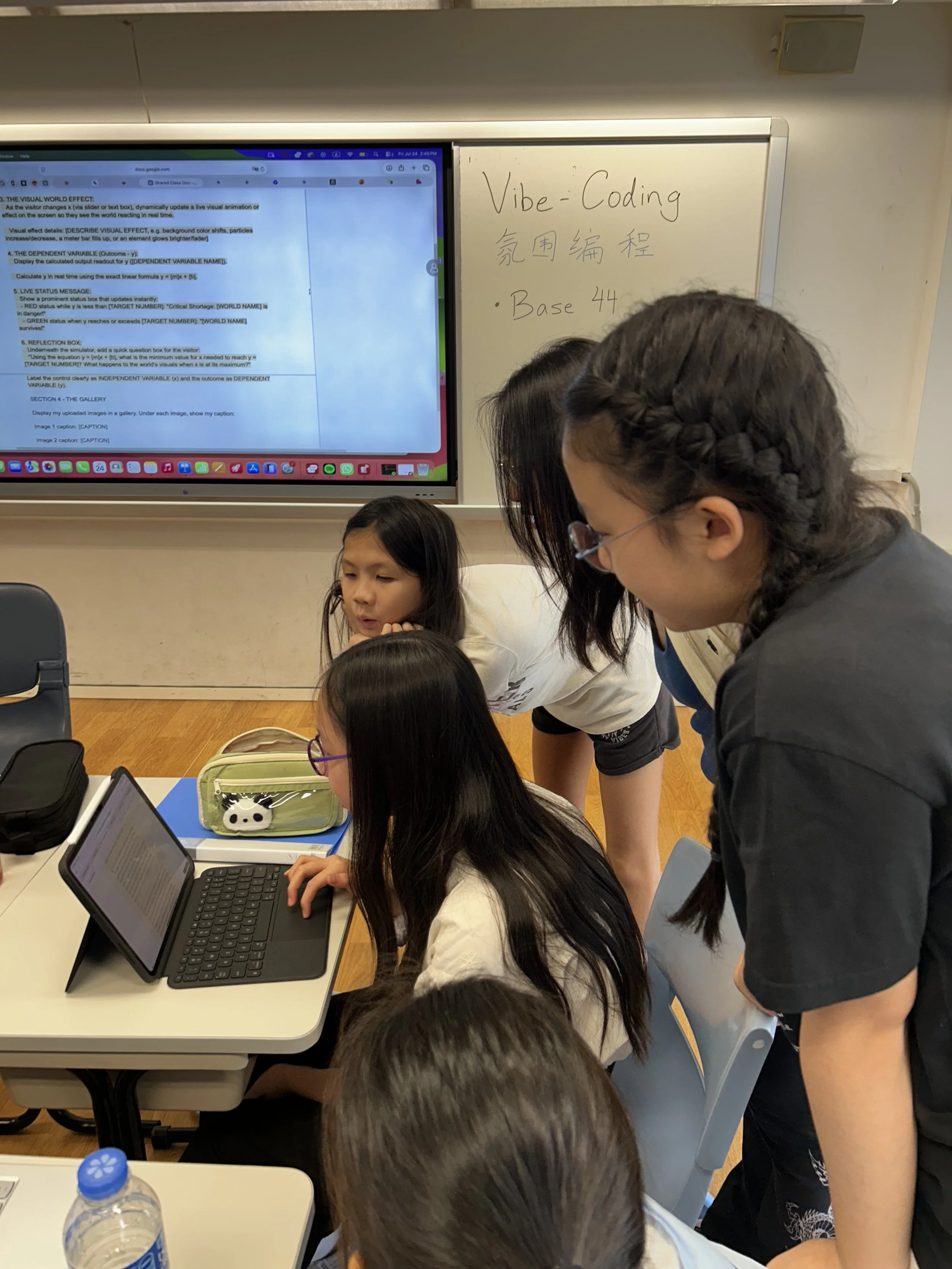 A classroom scene with students working on laptops at a table, one student is typing on a MacBook, while a teacher leans over to assist. There is a whiteboard with 'Vibe-Coding' written on it and some Chinese characters, as well as a large screen displaying instructions related to visual effects coding.