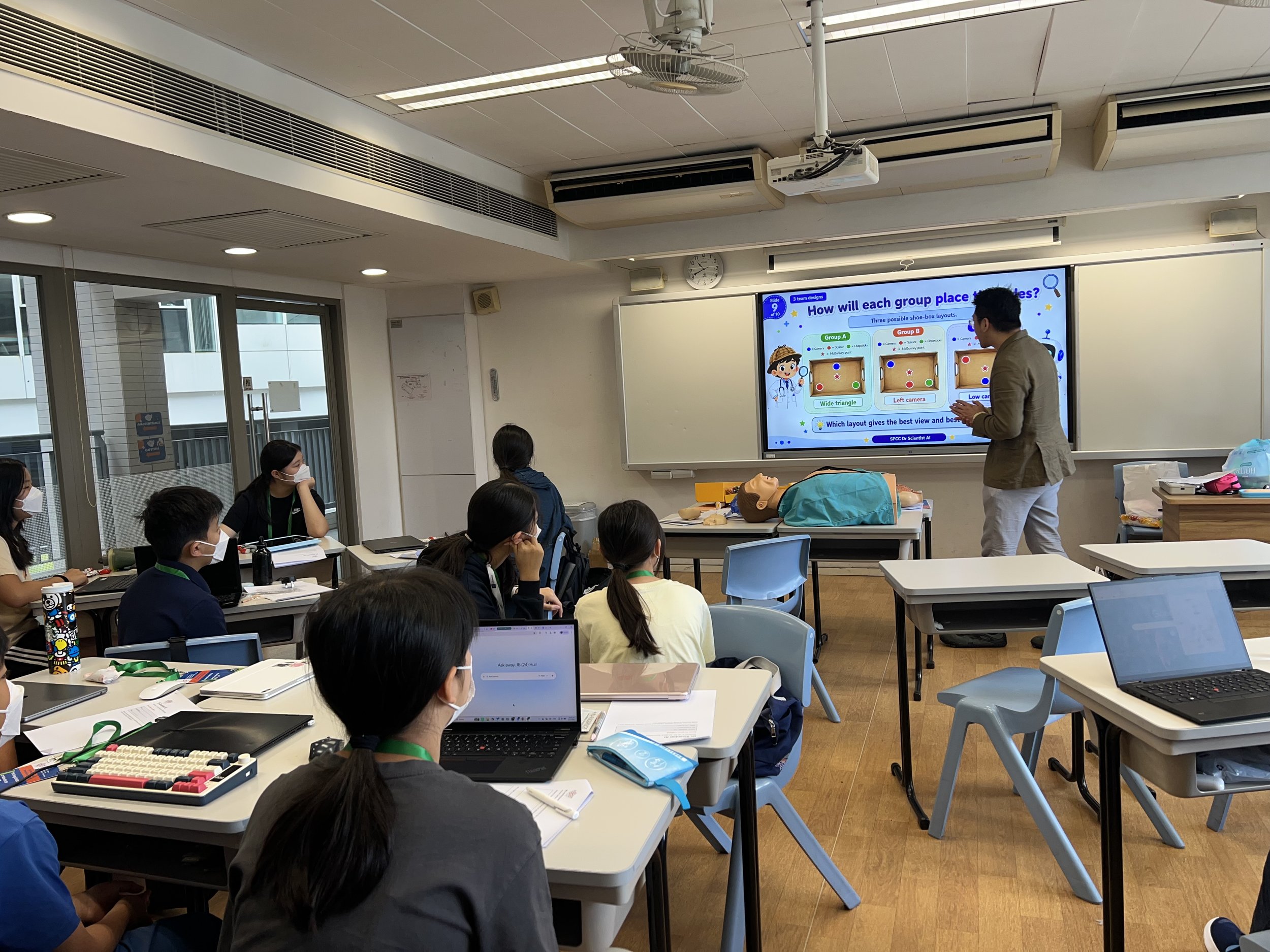 A classroom scene with students sitting at desks, some wearing masks. A teacher stands near a large screen presentation discussing different shoe-box layouts for a group activity, with medical training mannequins and medical supplies on a desk nearby.