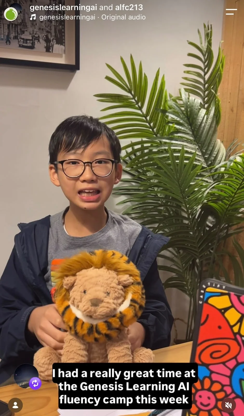 A boy with glasses holding a plush toy of a lion with a mane resembling a tiger's stripe pattern. He is sitting at a table with a colorful laptop and a large green potted plant behind him. There is a framed black-and-white photograph on the wall.