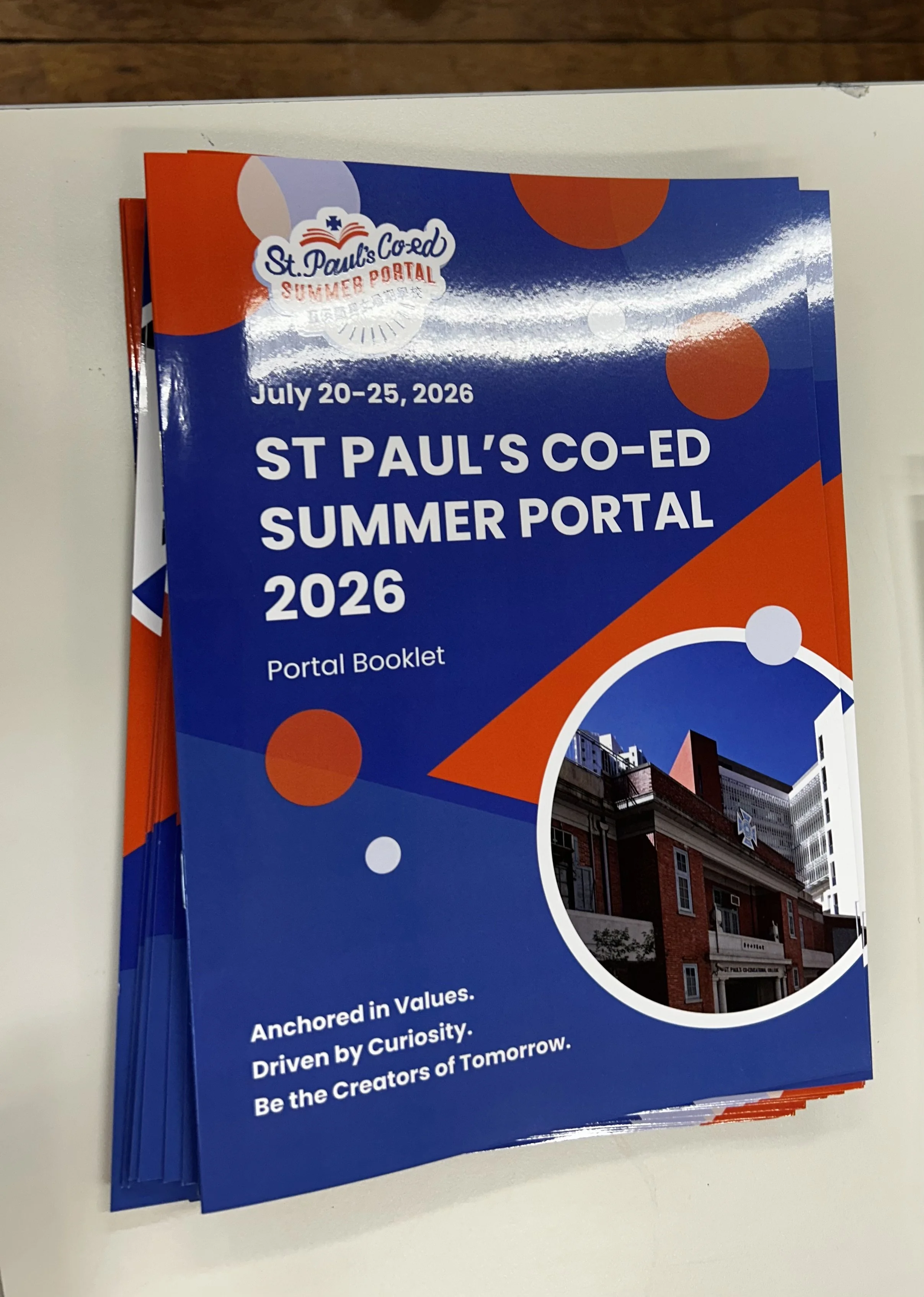 A stack of brochures for St. Paul's Co-Ed Summer Portal 2026, held from July 20-25, 2026. The brochure has a blue background with orange and white circles and includes a photograph of a brick building with white balconies and a star-shaped decoration.