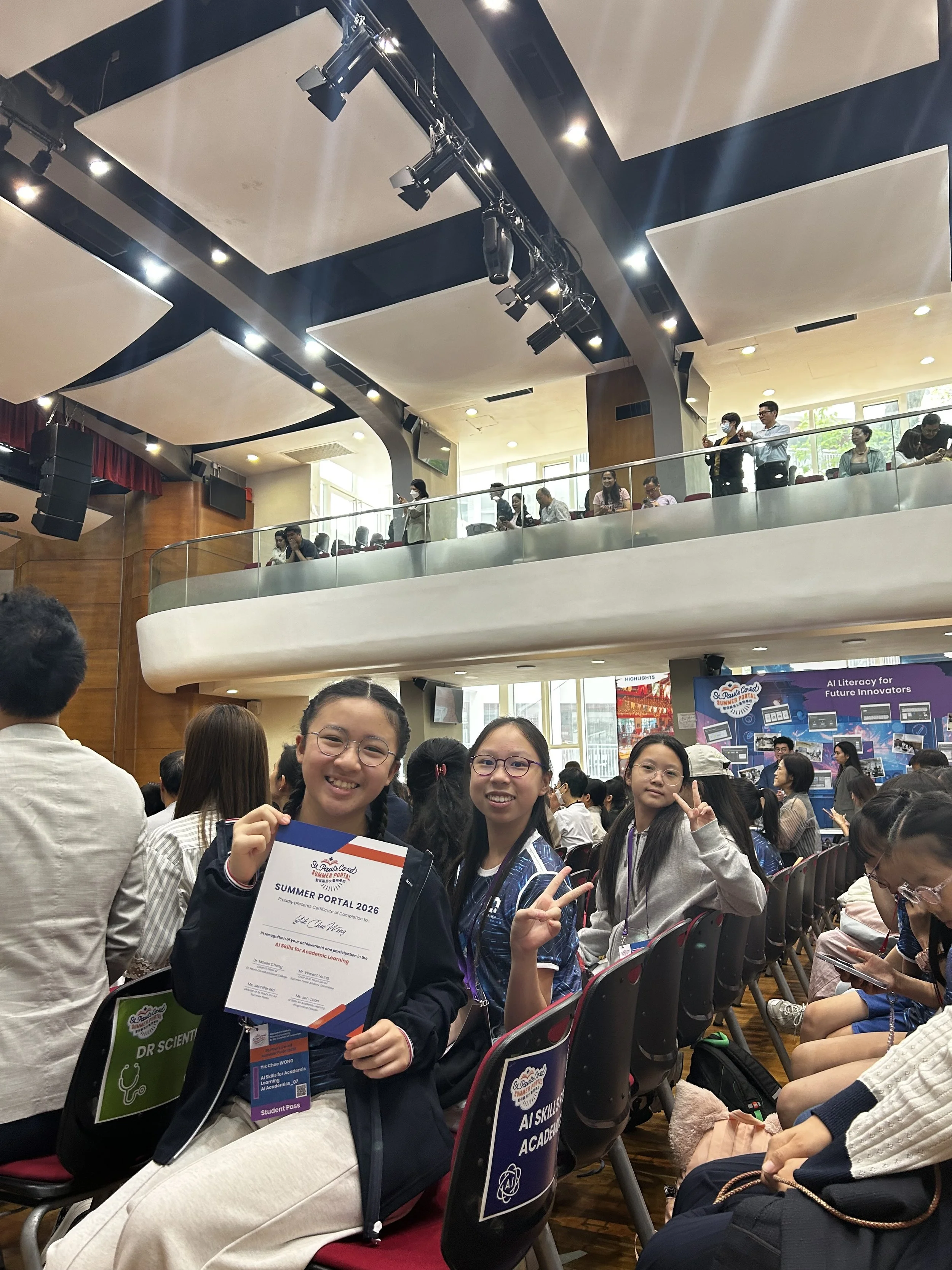 A group of students at an indoor event, some holding certificates and making peace signs, seated and standing in a large auditorium with a high ceiling and overhead lighting.