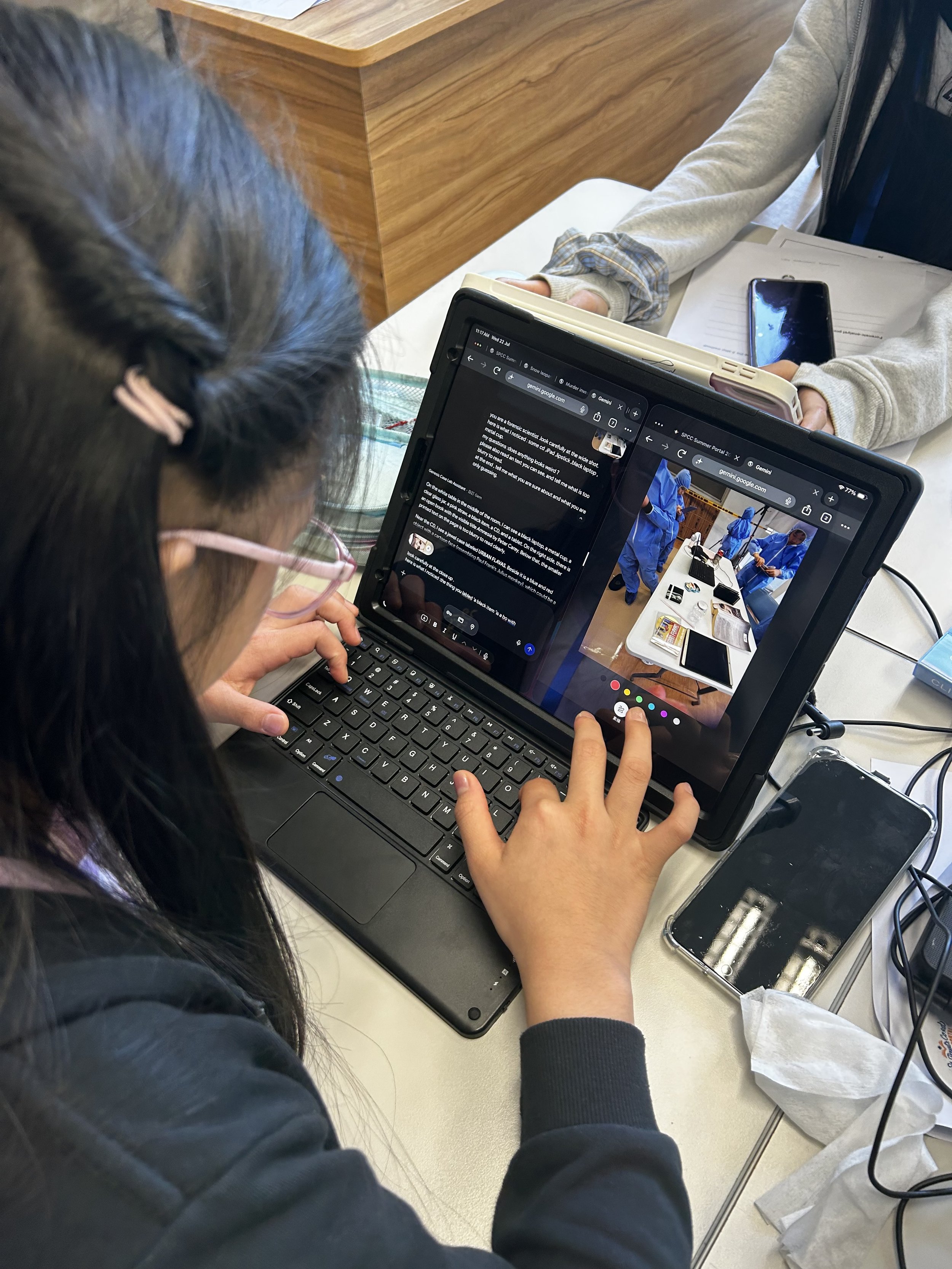 A person with long dark hair, wearing glasses and a black jacket, is using a laptop with multiple open windows and a tablet on top of the laptop. Another person with a light-colored hoodie and a scrunchie on their wrist is sitting across, holding a smartphone and some papers. The table also has various items including tissue, a black box with batteries, and wires.