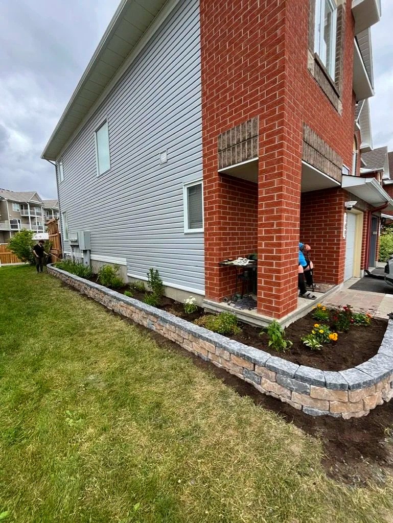View of a newly landscaped garden bed with a stone border around a house with vinyl siding, brick columns, and windows. Two people are working near the garden, one standing and one sitting.